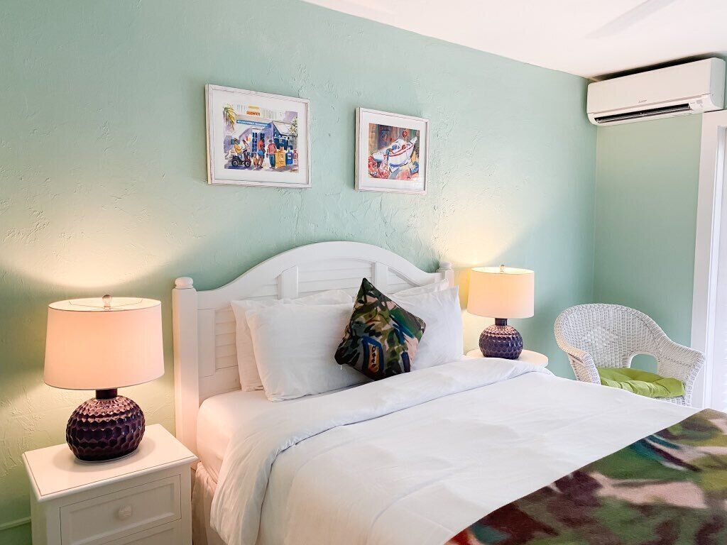 A neatly made bed with white linens, two framed pictures on the wall, a white nightstand with a lamp, and a wicker chair.