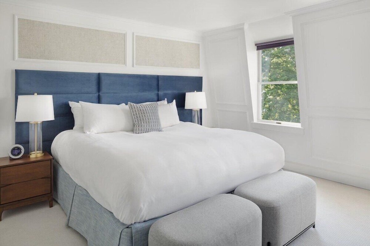 Un dormitorio con una cama grande, dos mesitas de noche con lámparas, una ventana con vistas y una mesita de noche con un reloj.