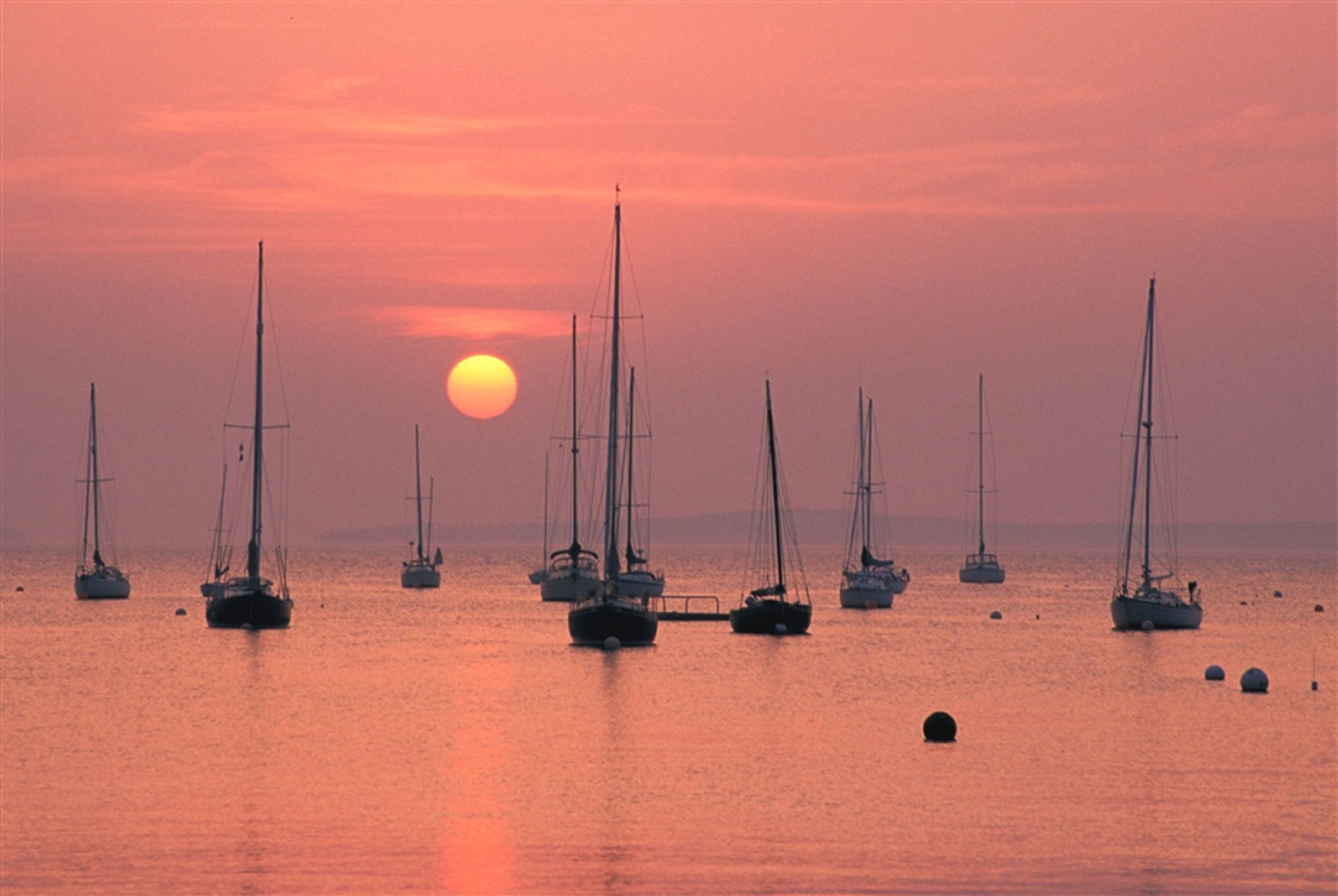 A sunset over a calm sea with several sailboats anchored.
