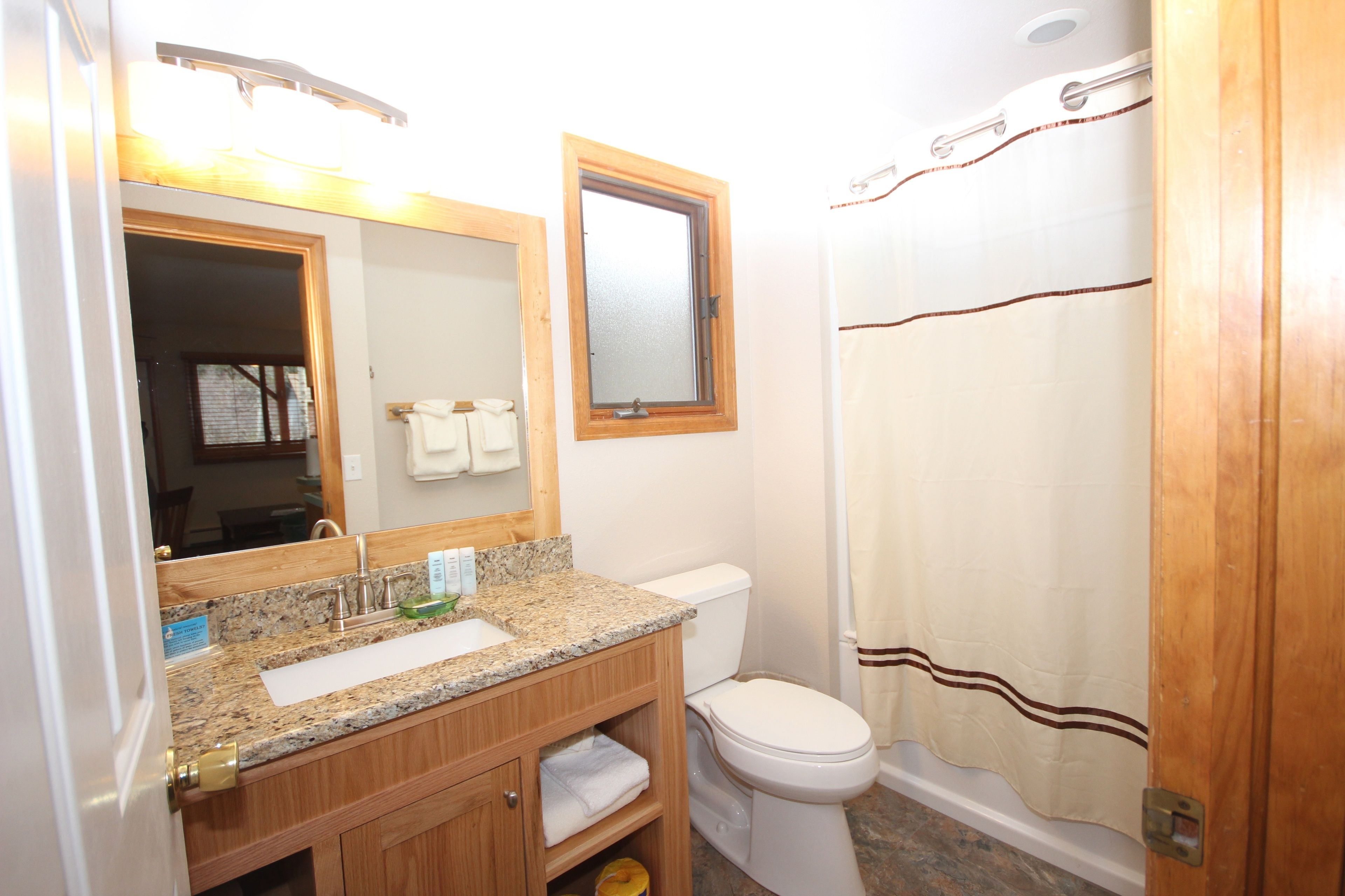 Un baño con lavamanos de encimera de granito, mueble de tocador de madera, inodoro blanco y cortina de ducha.