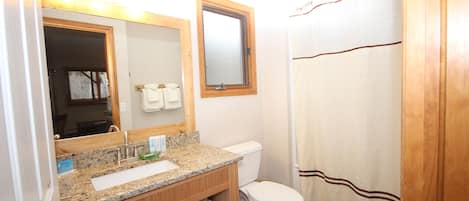 Un baño con lavamanos de encimera de granito, mueble de tocador de madera, inodoro blanco y cortina de ducha.