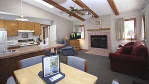 A modern kitchen with wooden cabinets, a dining area with a laptop on the table, a red sofa, and a fireplace.