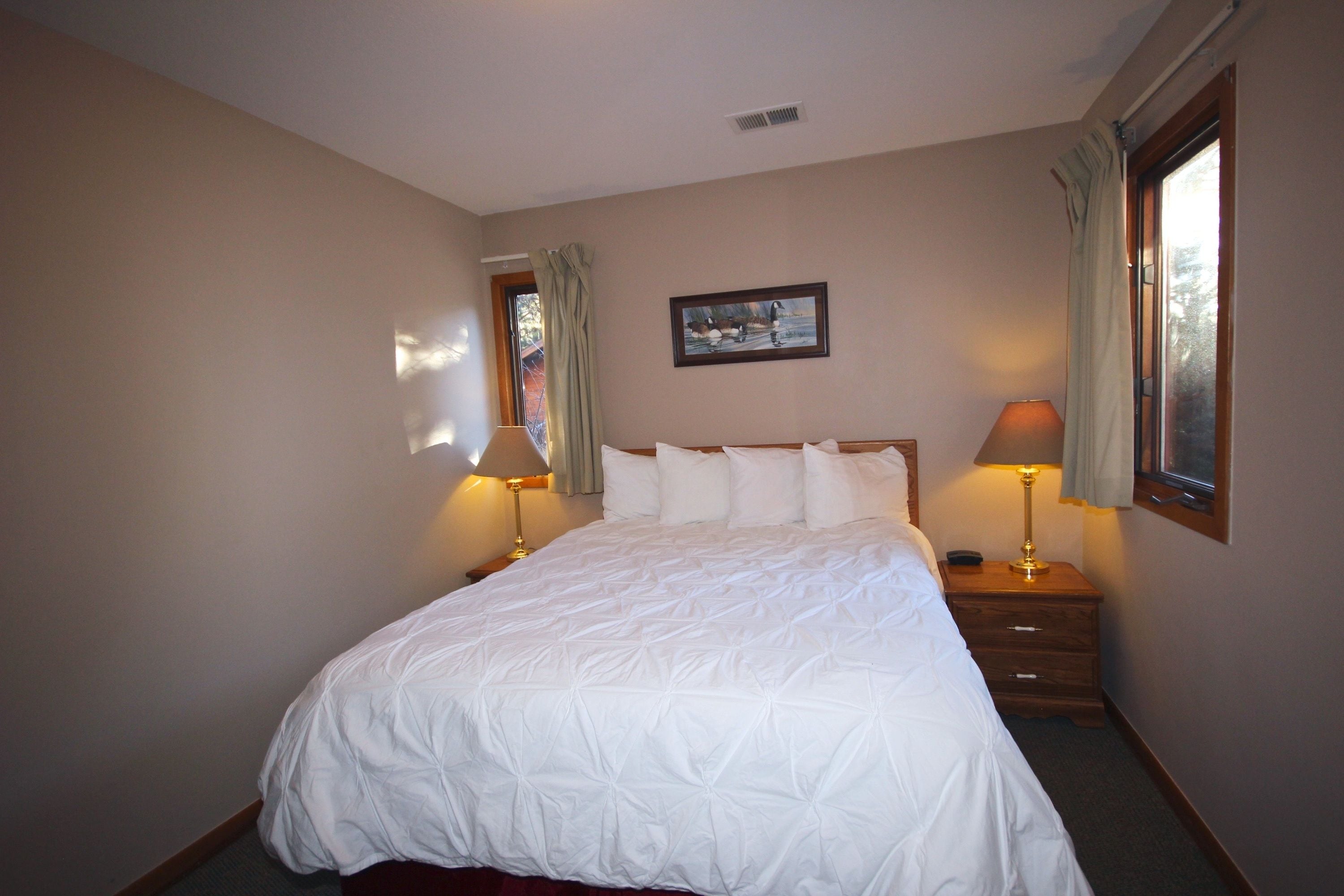 A neatly made bed with a white comforter in a room with beige walls, a framed picture on the wall, and two bedside lamps.