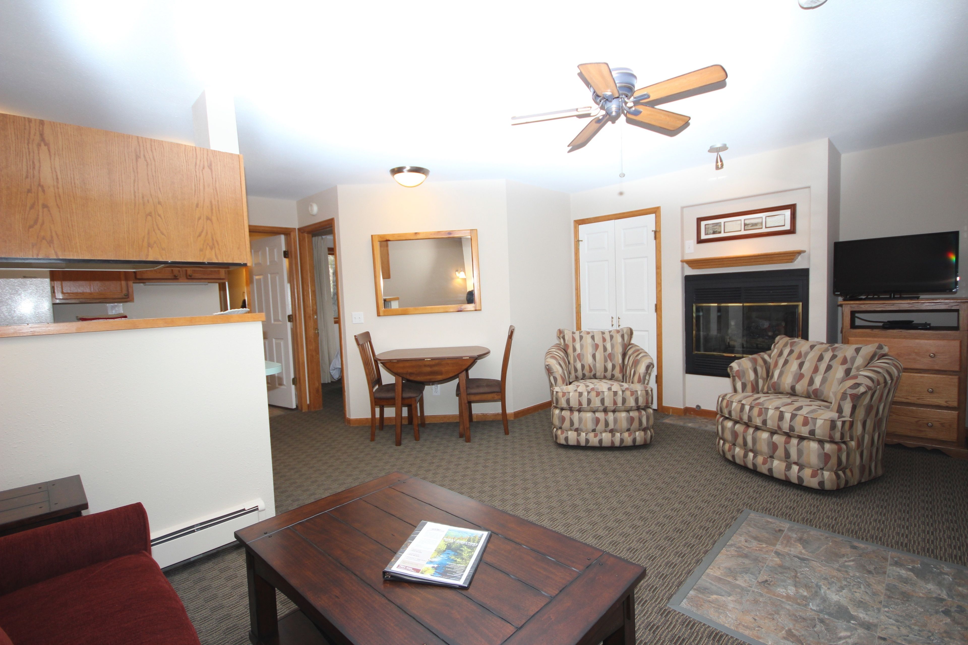 A living room with a wooden table, two armchairs, a ceiling fan, a fireplace, and a television.