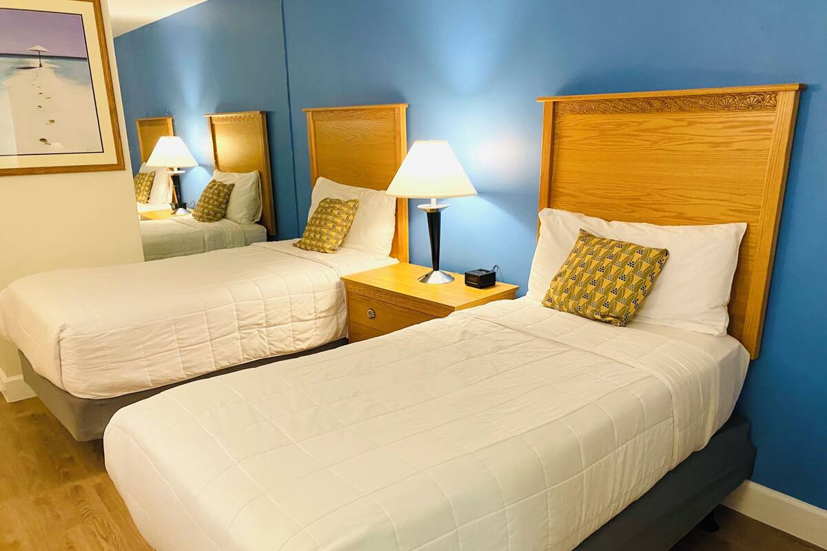 A hotel room with two single beds, each with a wooden headboard, a bedside table with a lamp, and a small nightstand.