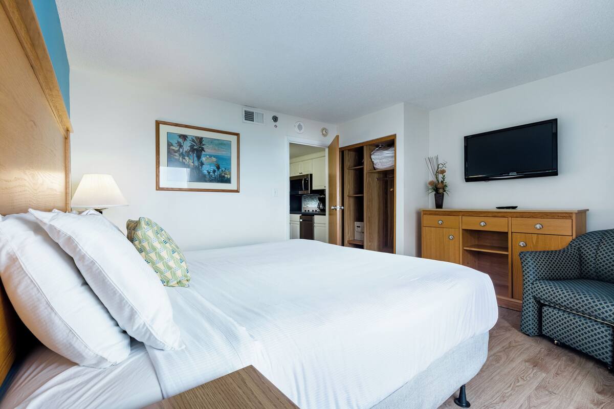 A hotel room with a large bed, a TV mounted on the wall, a wooden wardrobe, and a small desk.