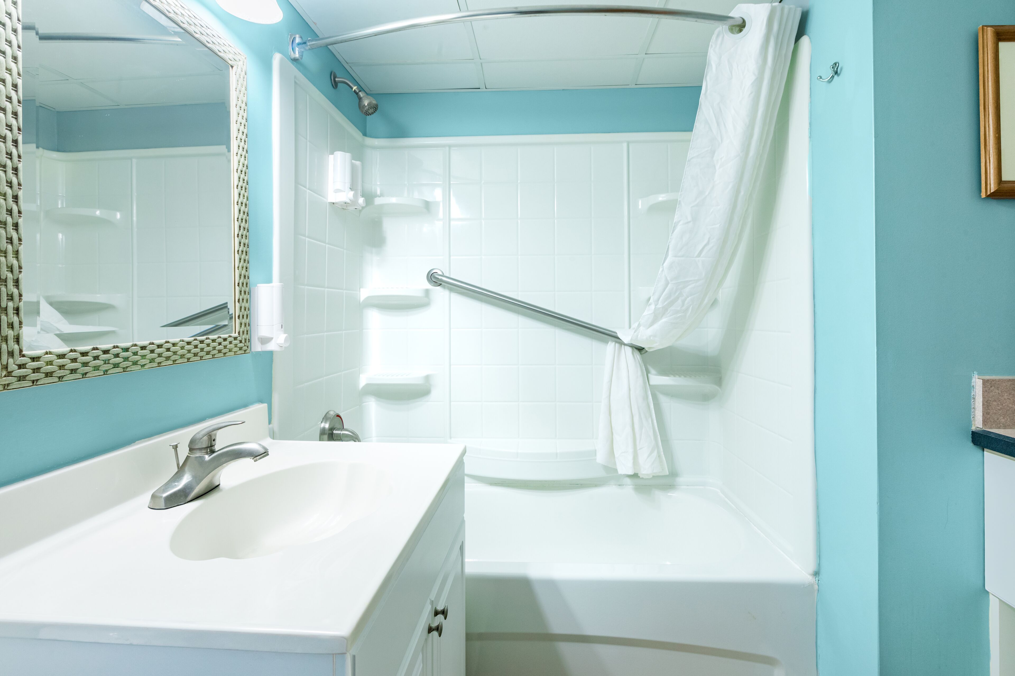 Une salle de bain avec un lavabo blanc, une baignoire, une douche équipée d’une barre d’appui et un miroir avec un cadre décoratif.