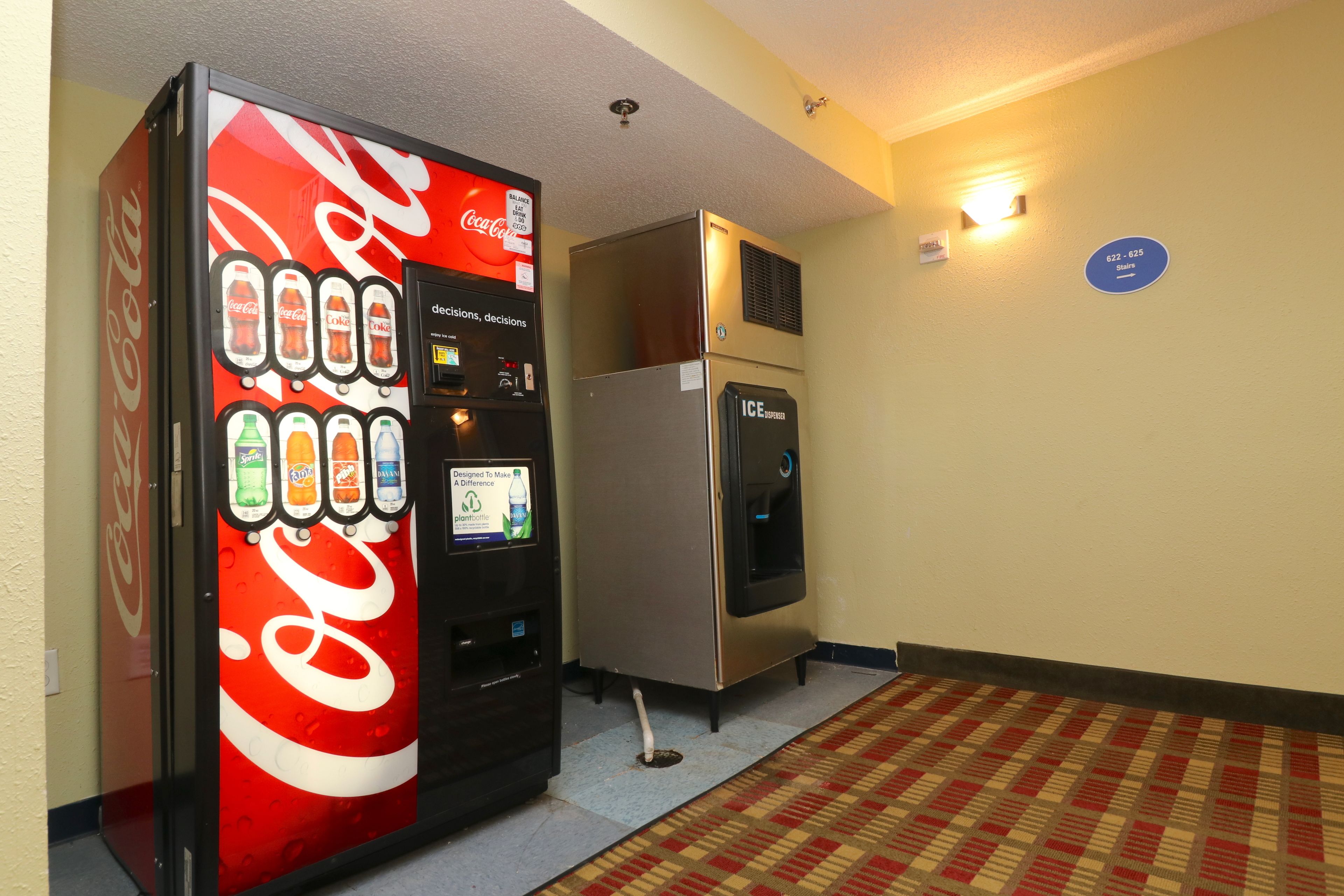 Máquina expendedora con Coca-Cola y otras bebidas, un refrigerador y suelo alfombrado en un pasillo.