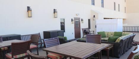 An outdoor patio with wooden tables and chairs, a sofa with cushions, and a white building in the background.