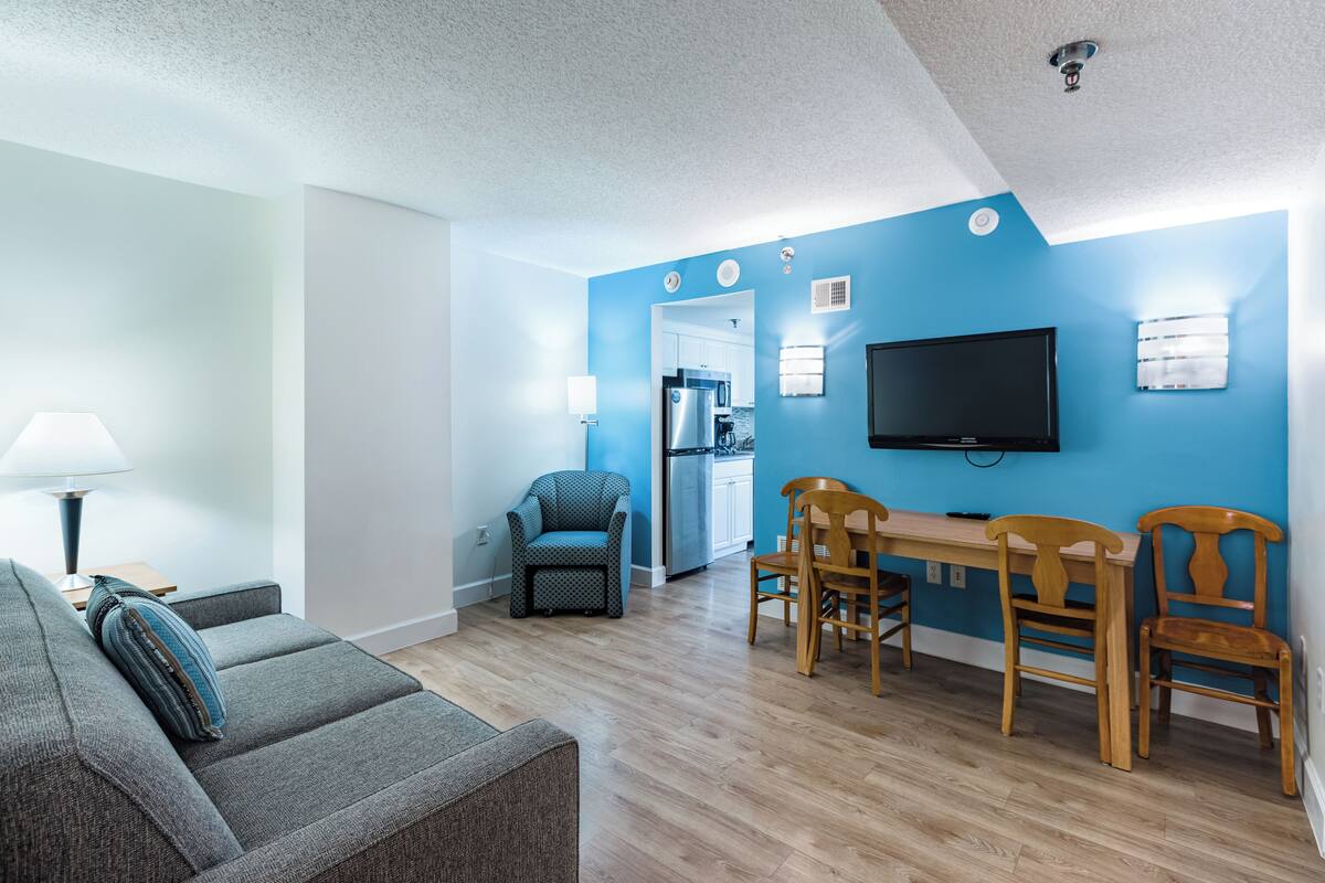A modern living room with a grey sofa, a dining table with wooden chairs, a blue wall, and a flat-screen TV mounted above the table.