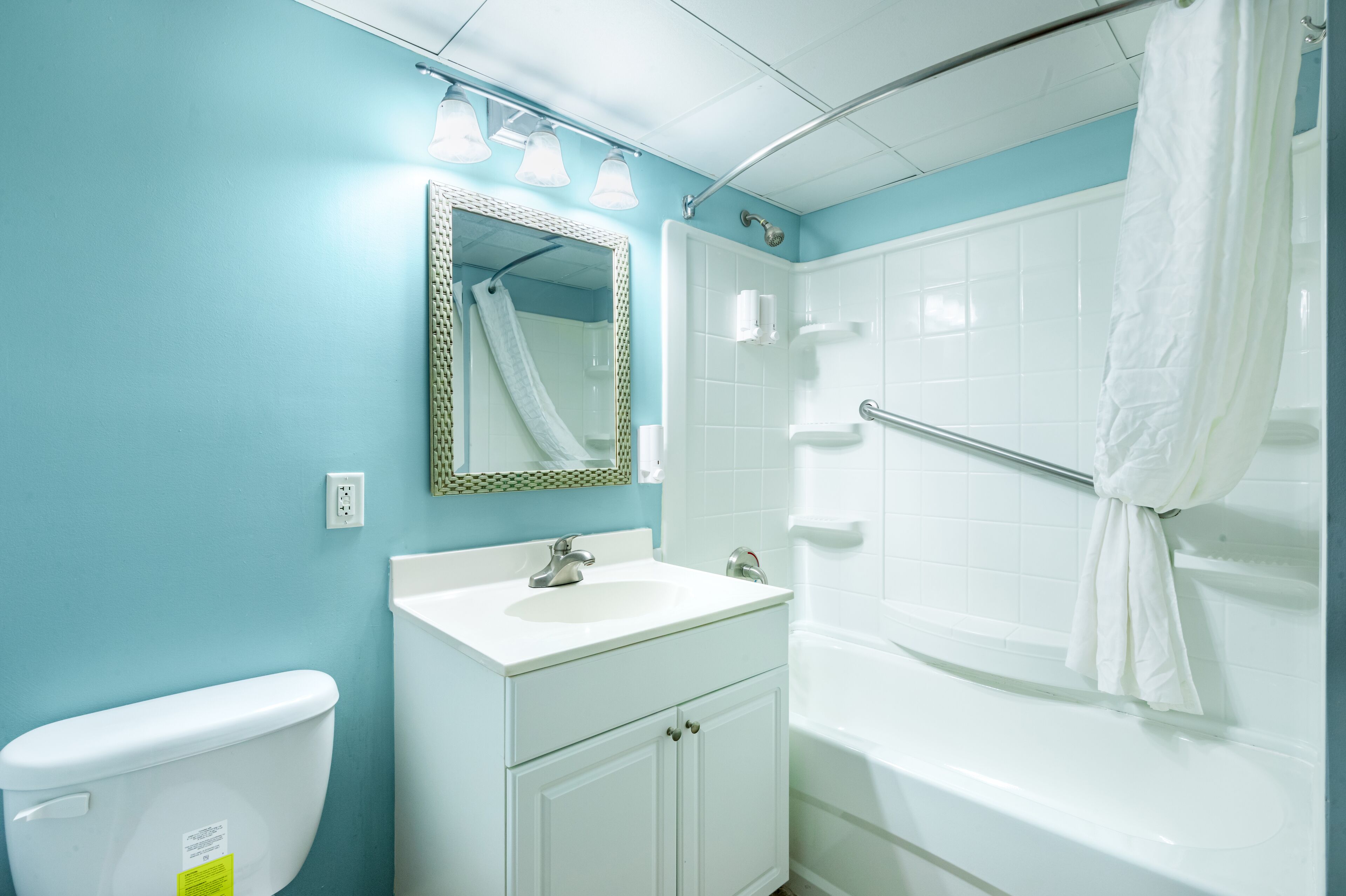 Ein Badezimmer mit einem weißen Waschbecken, einer Toilette, einer Dusche mit weißem Vorhang und einem Spiegel mit verzierter Rahmen.