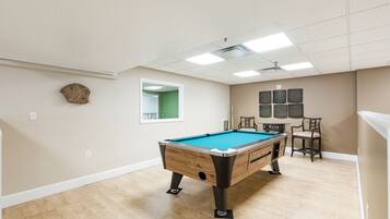 A pool table in a room with a wooden floor, a ceiling fan, and a window with a green view.