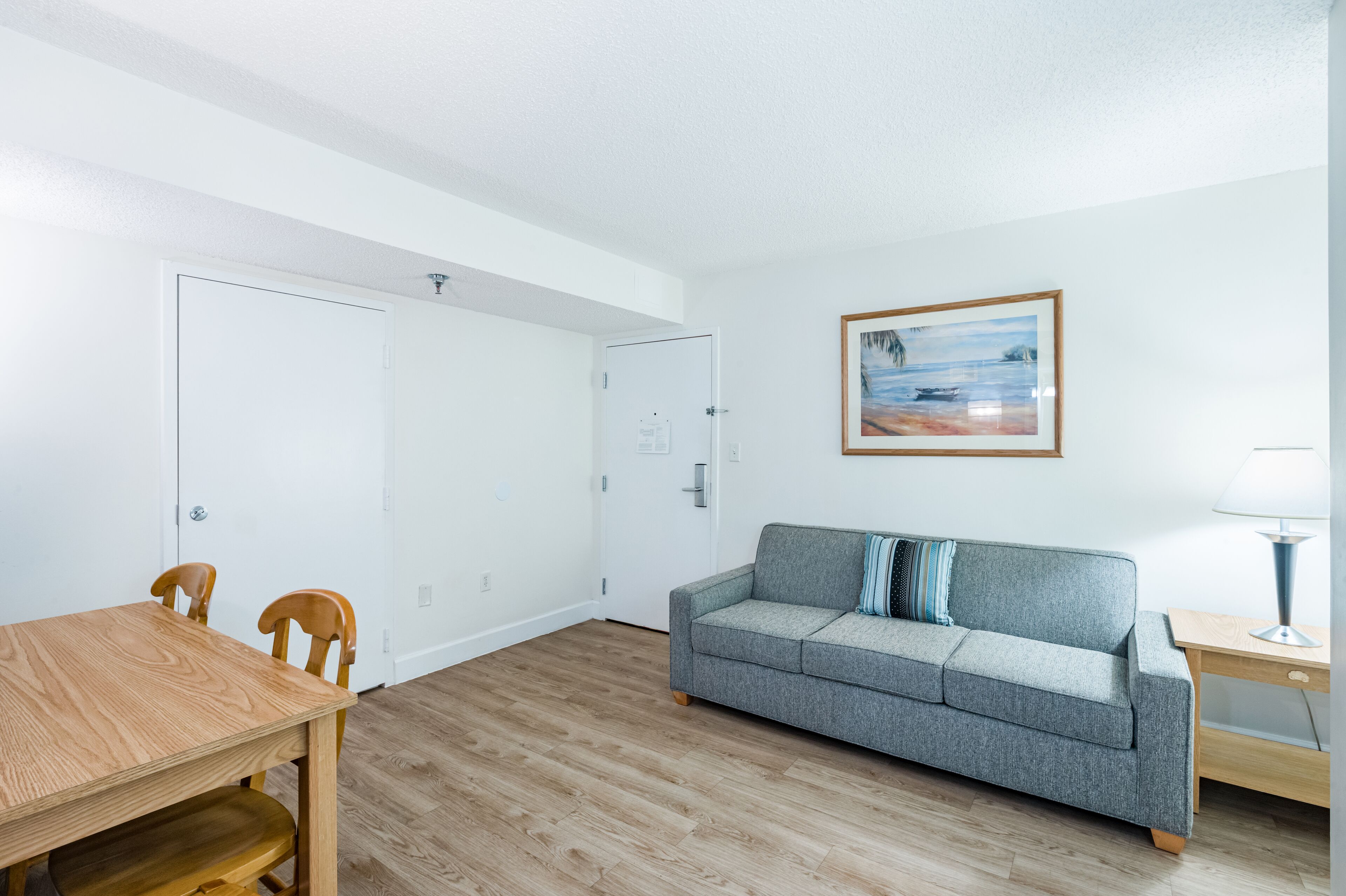 A room with a wooden dining table, two wooden chairs, a grey sofa with a blue and white striped pillow, a wooden side table with a lamp, and a painting of a beach scene on the wall.