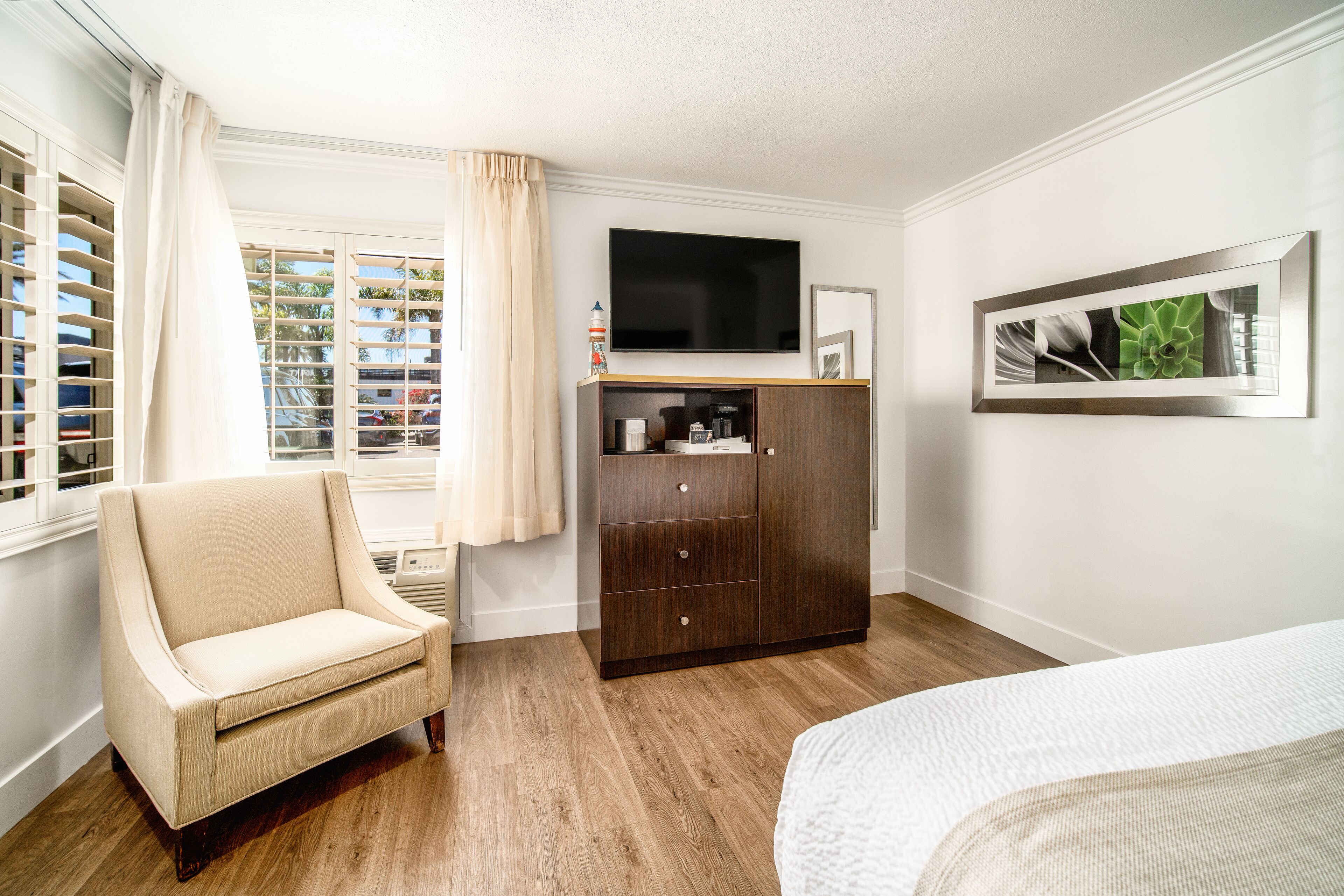 A bedroom with a wooden floor, a beige armchair, a dark wooden wardrobe, a bed with white bedding, a flat-screen TV mounted on the wall, and a framed picture above the TV.