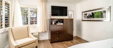 A bedroom with a wooden floor, a beige armchair, a dark wooden wardrobe, a bed with white bedding, a flat-screen TV mounted on the wall, and a framed picture above the TV.