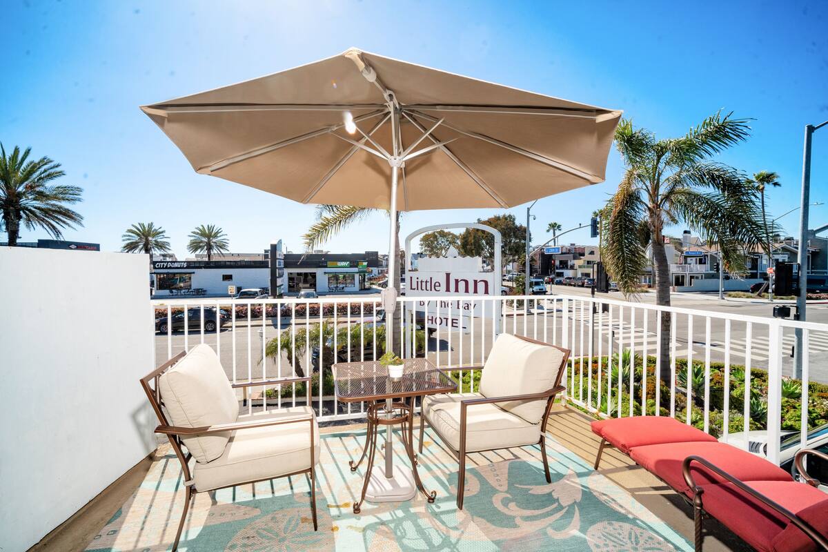 Un balcon avec une table et des chaises, un grand parasol et une vue sur la rue, avec des palmiers et une enseigne indiquant la présence d’un petit hôtel.