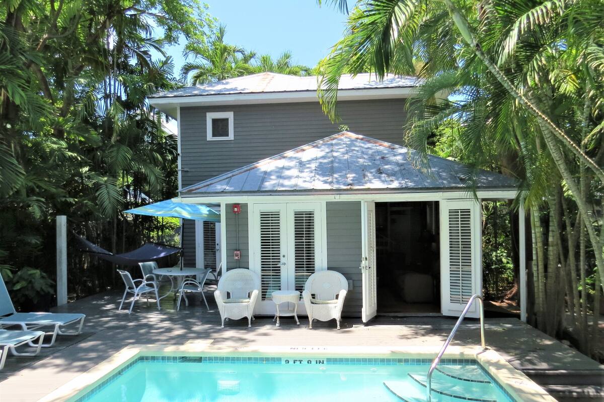 A pool area with a covered seating area, lounge chairs, and a pool with a depth marker.