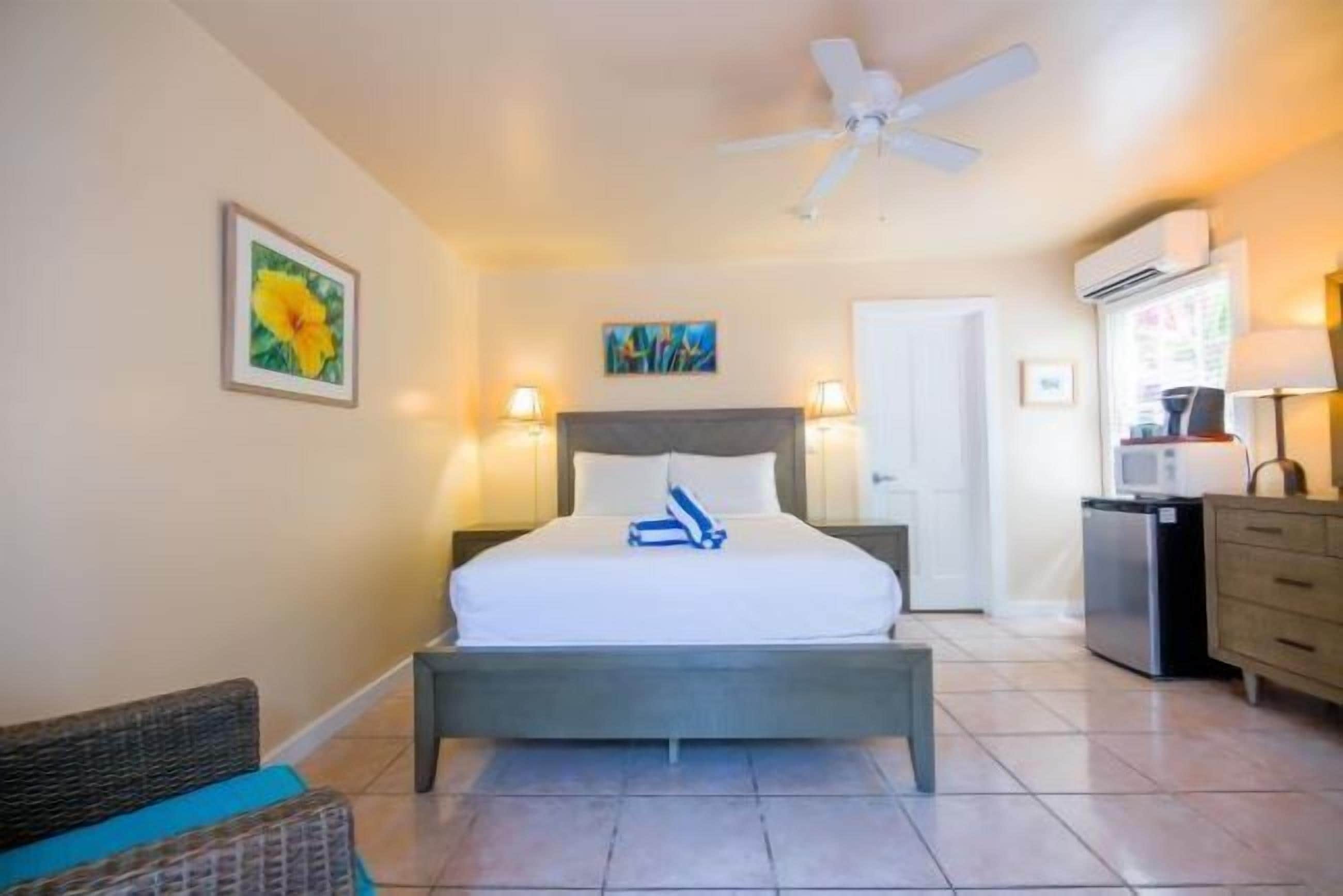 Une chambre d’hôtel avec un lit, un ventilateur de plafond, une petite kitchenette et une porte donnant sur une autre pièce.