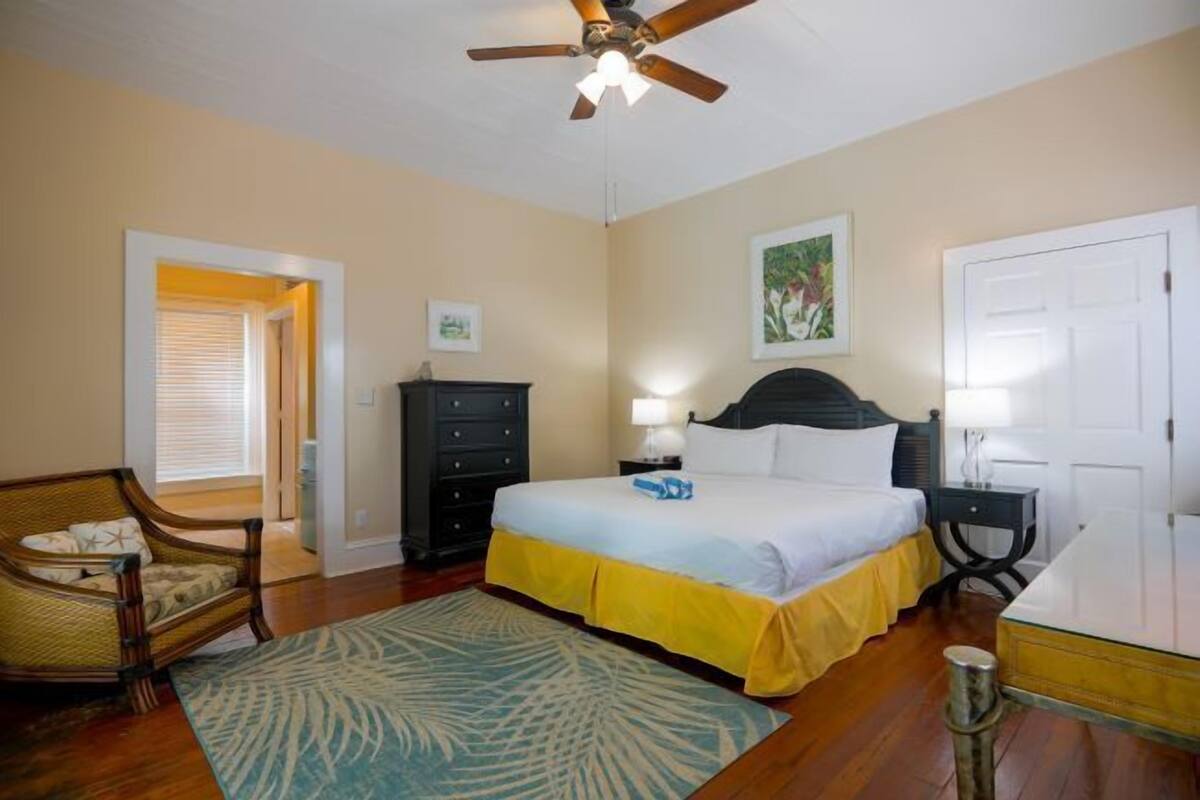 A bedroom with a bed, a chair, a dresser, a nightstand, a ceiling fan, and a door.