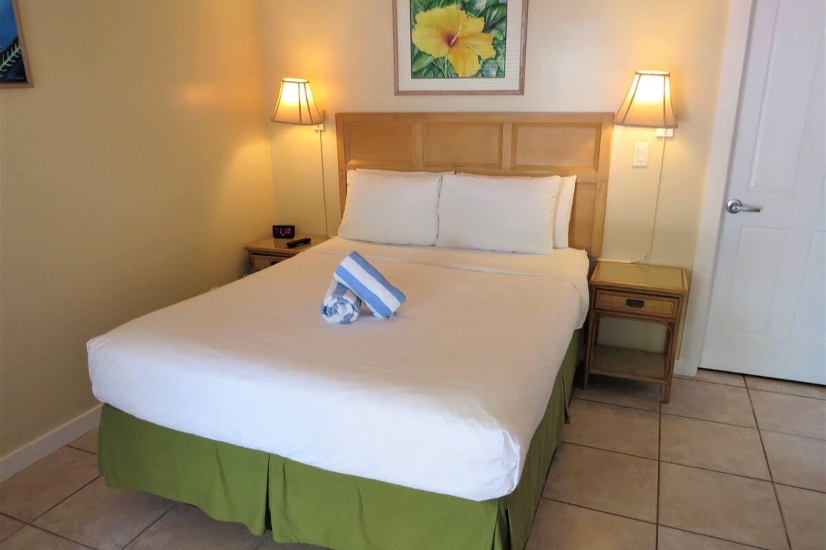 A neatly made bed with a green skirt, two bedside tables with lamps, a clock, and a floral painting on the wall.