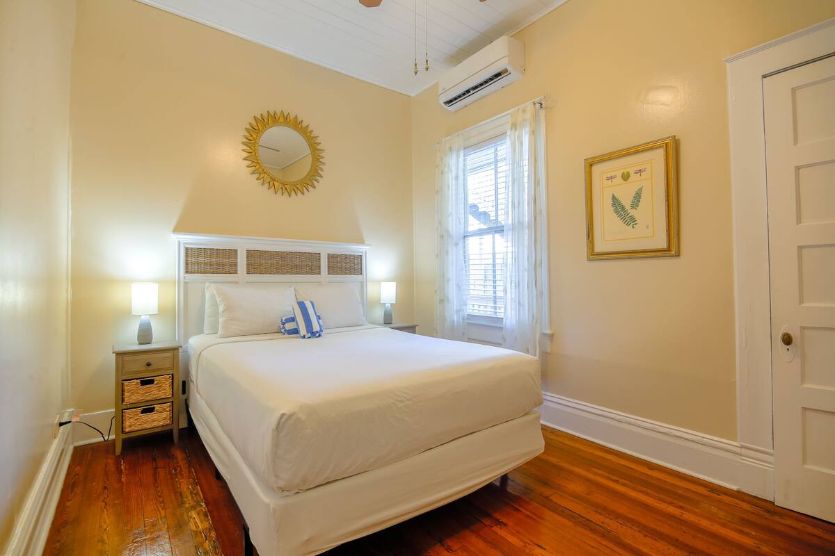 A bedroom with a large bed, a nightstand, a wall-mounted mirror, and a framed botanical print.
