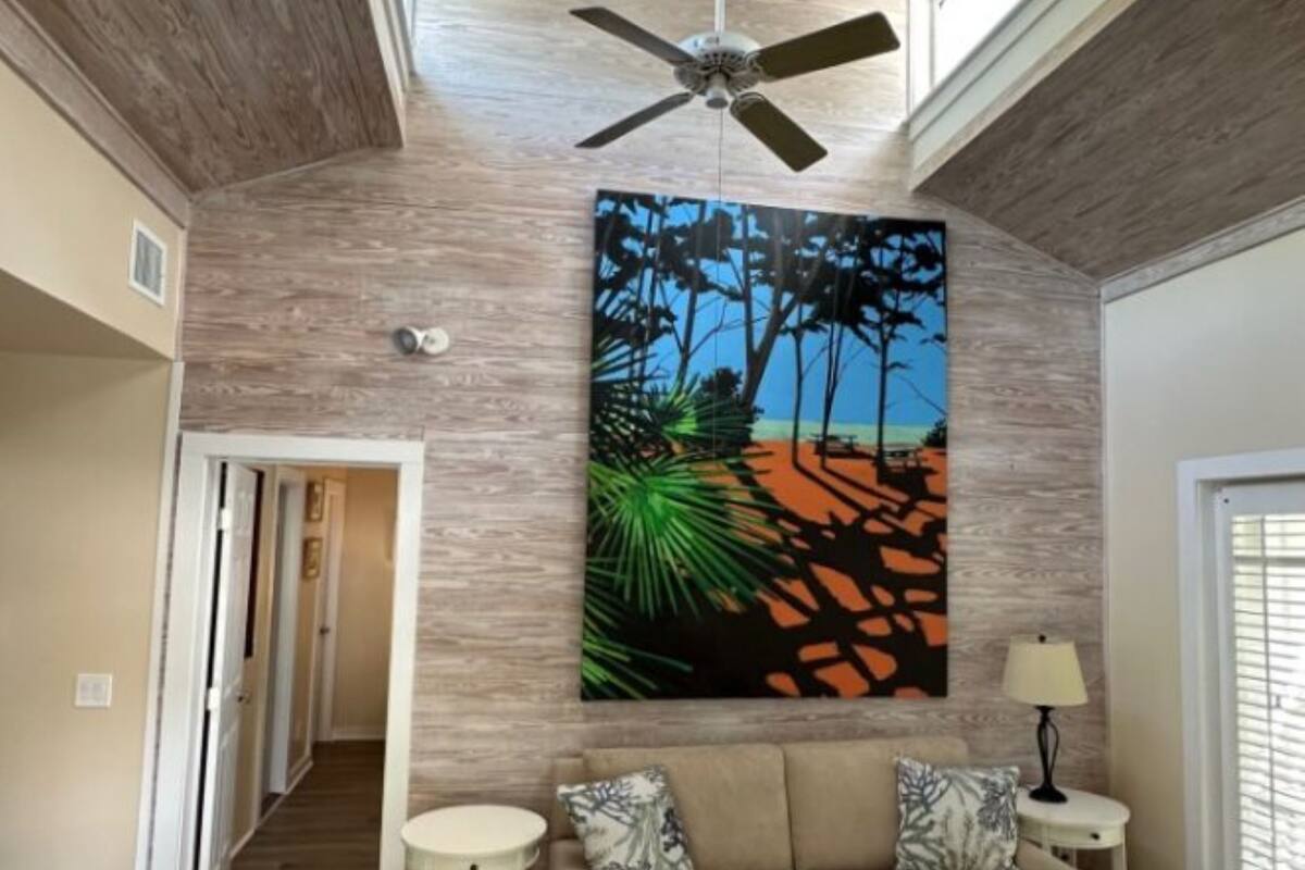 A spacious living room with a high ceiling, a ceiling fan, a large abstract painting, a beige sofa, two side tables, and a view of a hallway.
