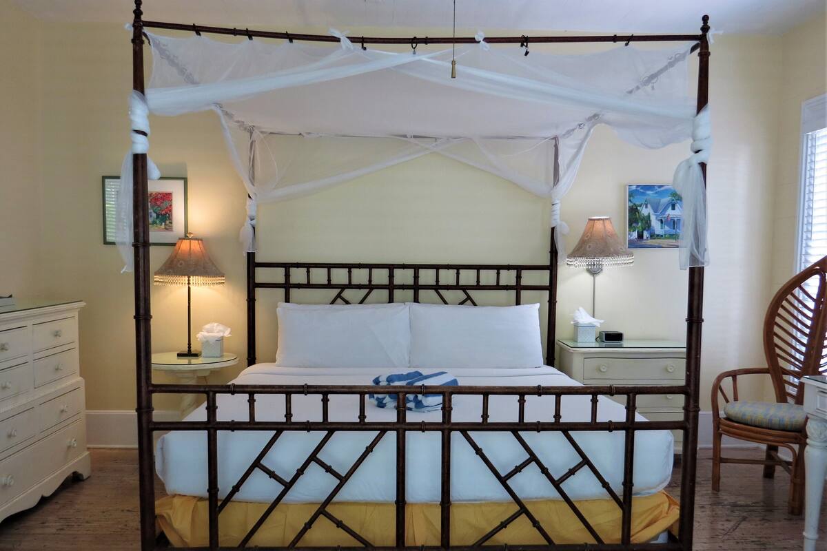 A four-poster canopy bed with a wooden frame, two bedside tables, a chair, and a dresser.