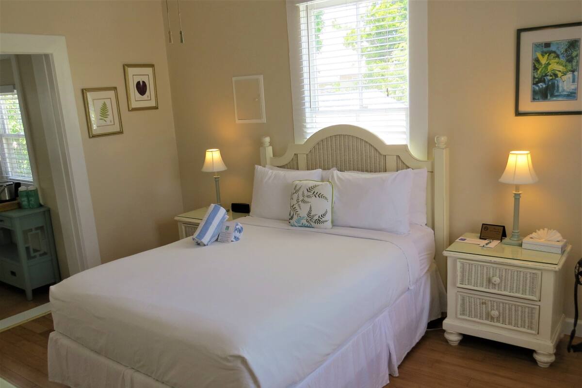 A neatly made bed with white linens, two bedside tables with lamps, a small nightstand, and a window with blinds.