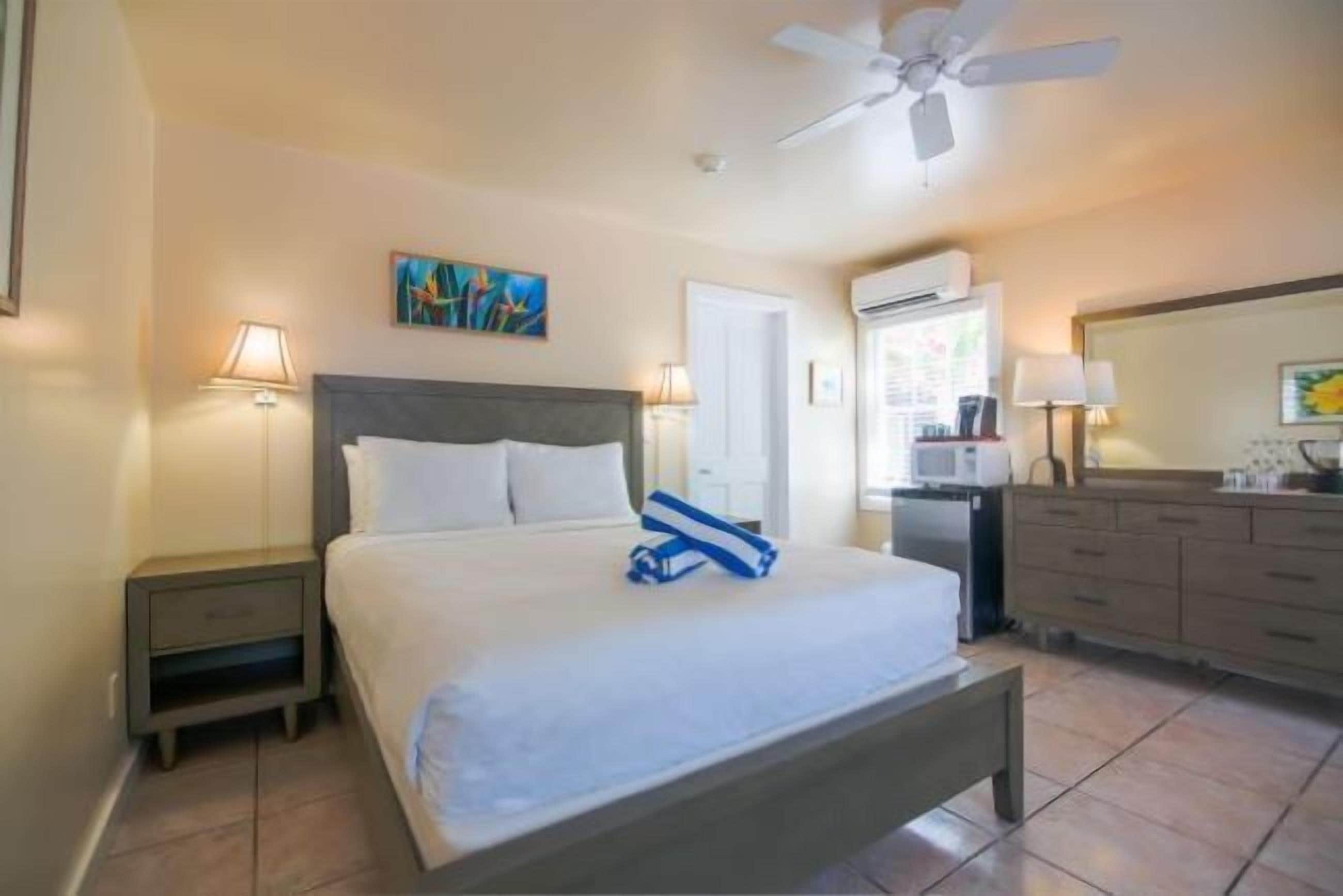 A hotel room with a bed, a nightstand, a desk, a chair, a ceiling fan, a painting, and a bathroom with a mirror and sink.