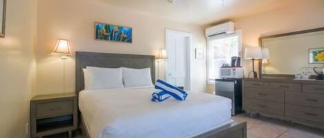 Camera d'albergo con un letto, un comodino, una scrivania, una sedia, una ventola a soffitto, un quadro e un bagno con uno specchio e un lavandino.