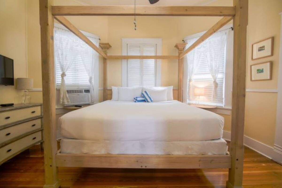 A four-poster bed in a room with wooden beams, white bedding, a television on a dresser, and a window with blinds.