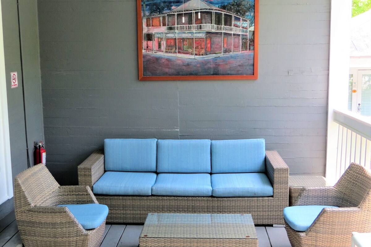 A covered porch with a blue sofa, two wicker chairs, and a matching ottoman. A ceiling fan and a framed painting are visible on the wall.