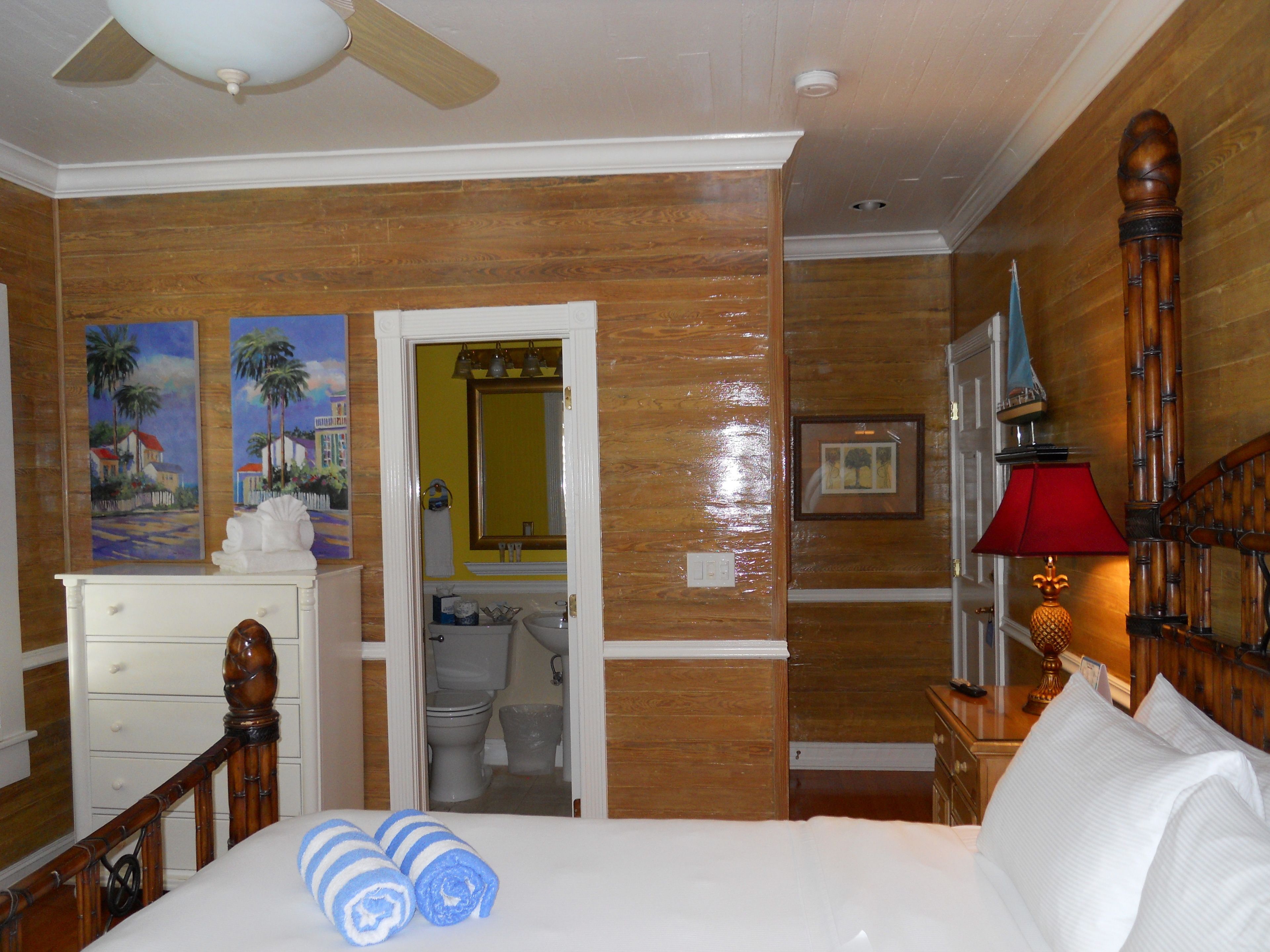 Habitación de hotel con una cama, cabecera de madera, mesita de noche con lámpara, un cuadro en la pared y un baño visible a través de una puerta abierta.