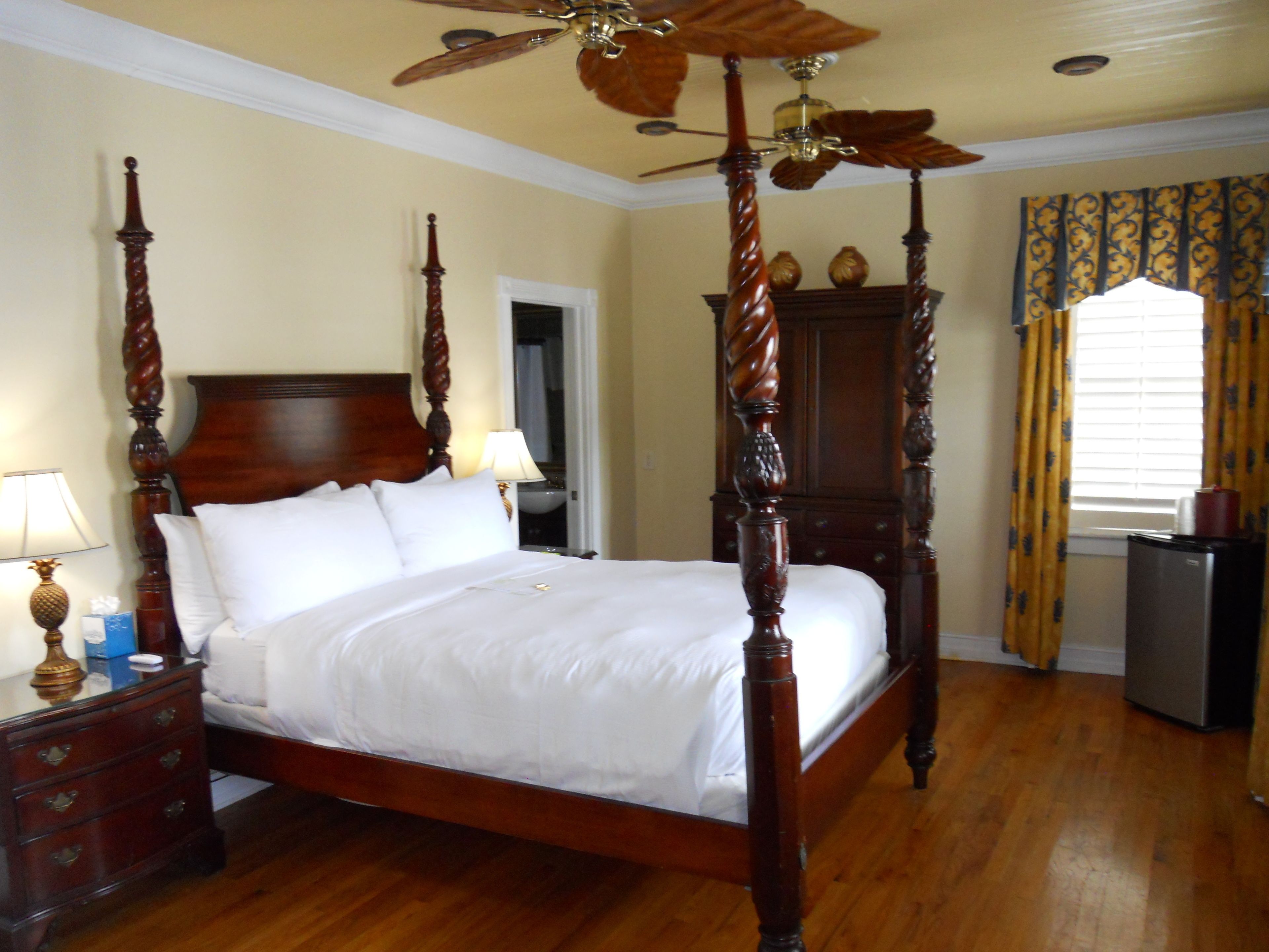 A four-poster bed with a wooden headboard and footboard, a ceiling fan, a nightstand with a lamp, and a dresser.