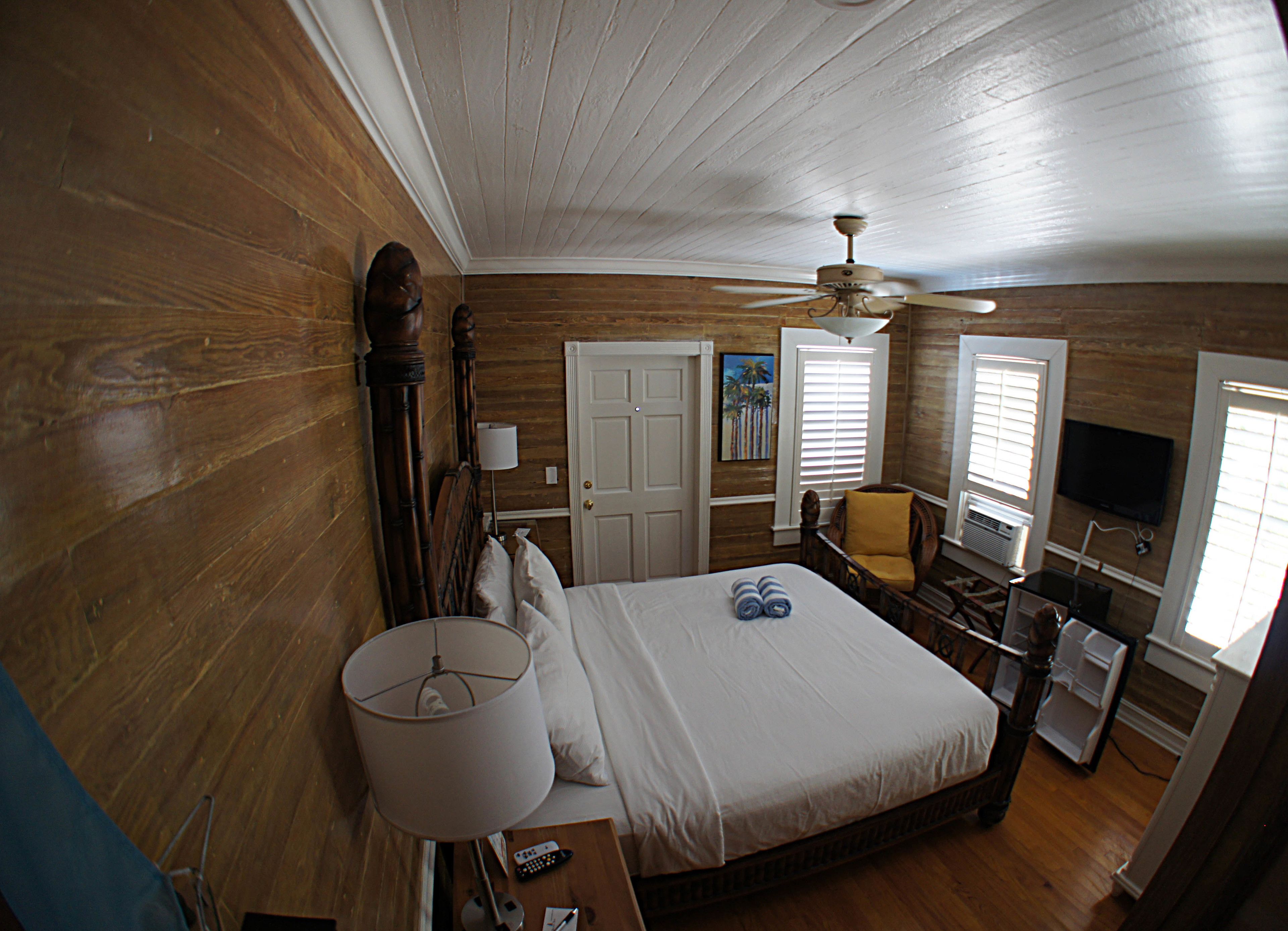 Une chambre avec un plancher en bois, un lit avec du linge de lit blanc, un ventilateur de plafond, une chaise, un téléviseur et une fenêtre avec des stores.