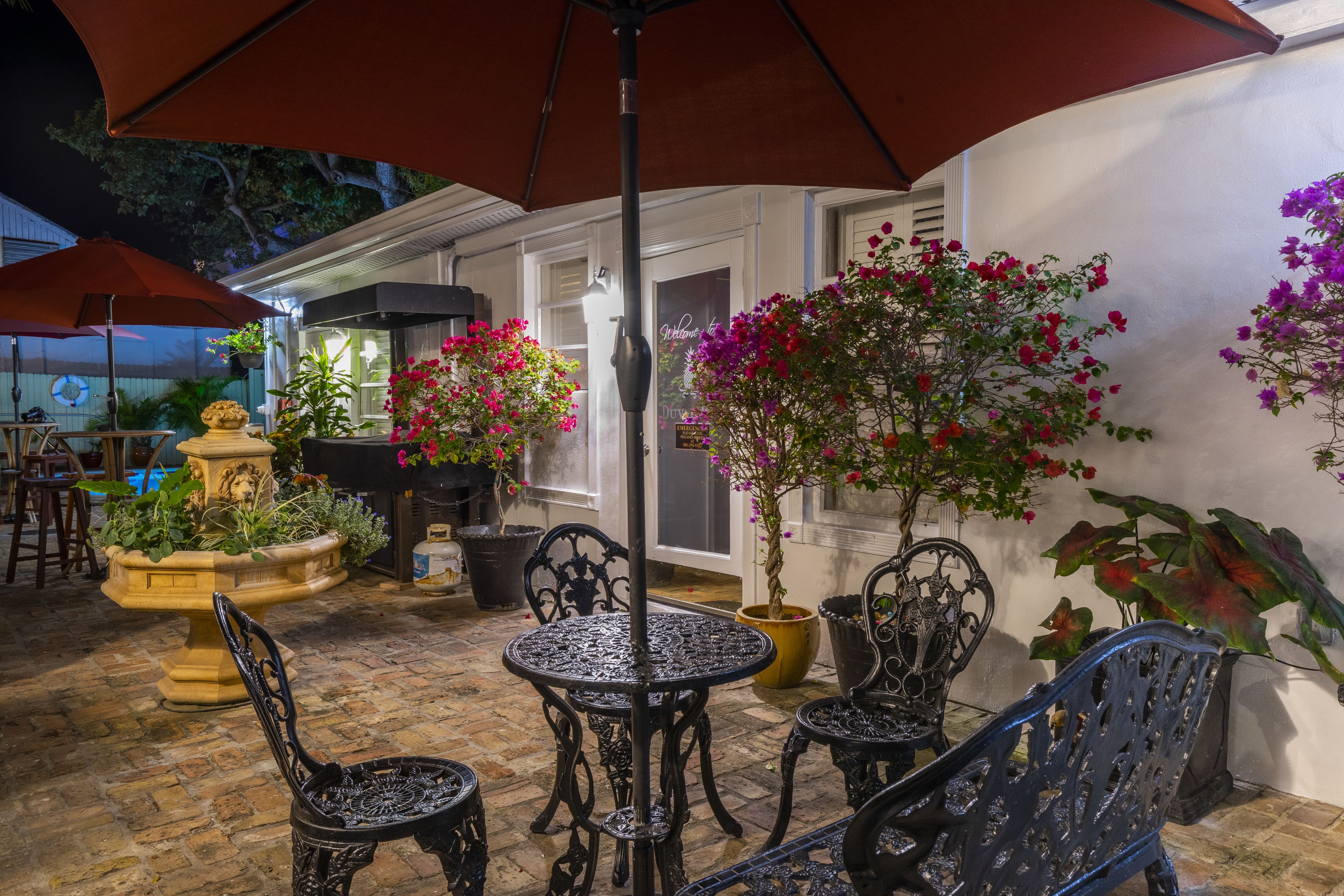 An outdoor patio with a round table and four chairs, surrounded by potted plants and a large umbrella.