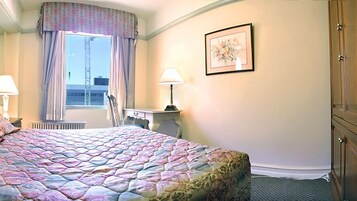 Habitación de hotel con una cama grande, un escritorio, una silla, una ventana con cortinas, un radiador y un cuadro en la pared.