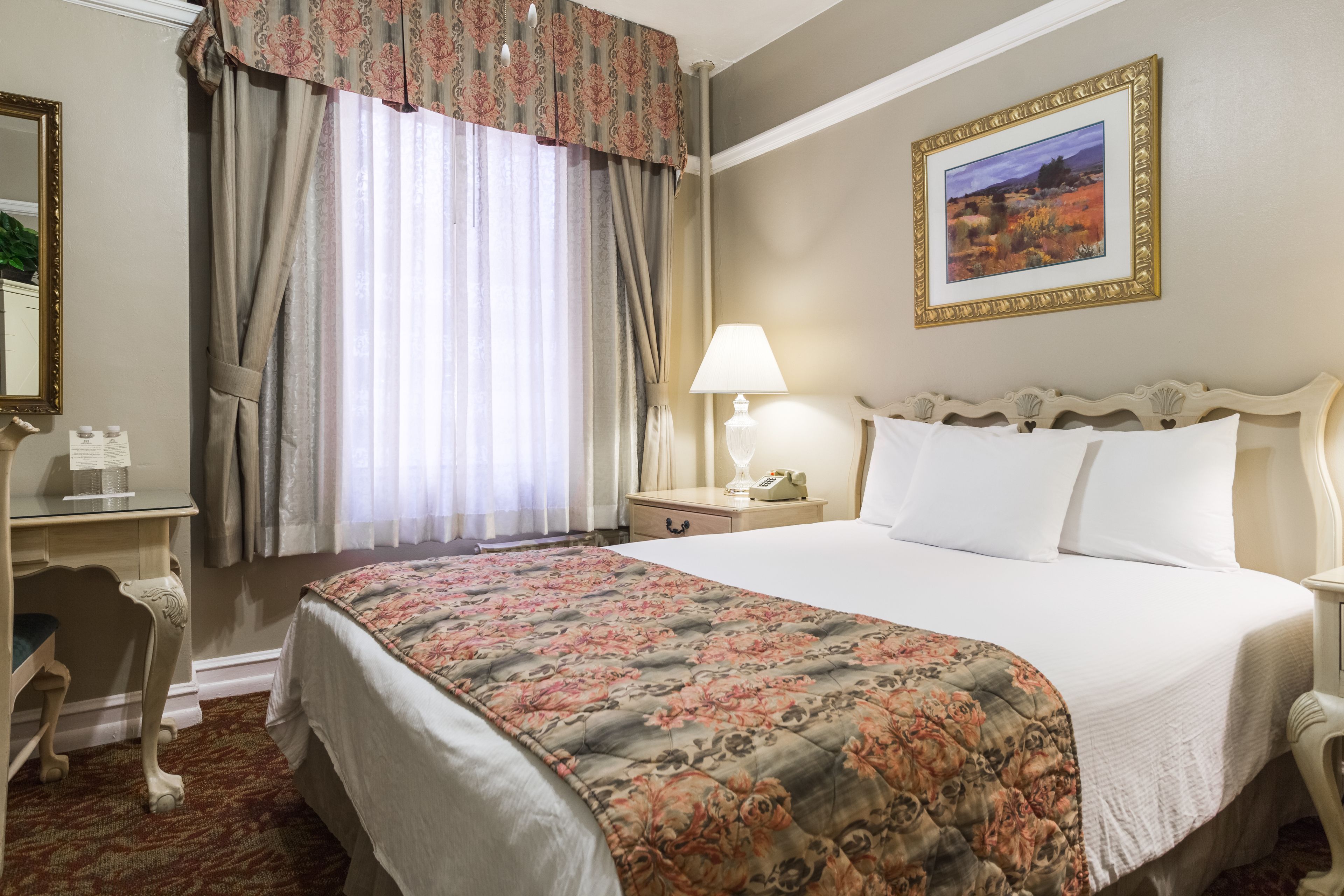 Habitación de hotel con una cama grande, un escritorio con una silla, una mesita de noche con lámpara y un cuadro en la pared.