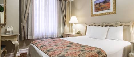 A hotel room with a large bed, a desk with a chair, a bedside table with a lamp, and a framed picture on the wall.