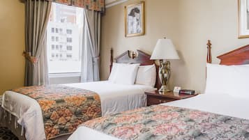 Habitación de hotel con dos camas, una mesita de noche con lámpara, un cuadro enmarcado en la pared y una ventana con cortinas.