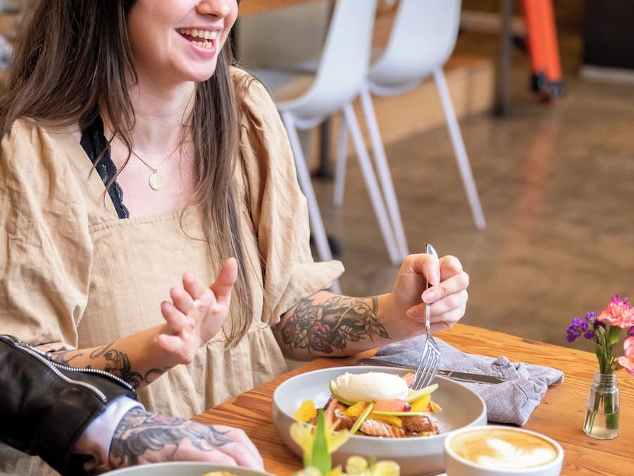Una persona sentada en una mesa de madera con platos de comida, una taza de café y un florero con flores.