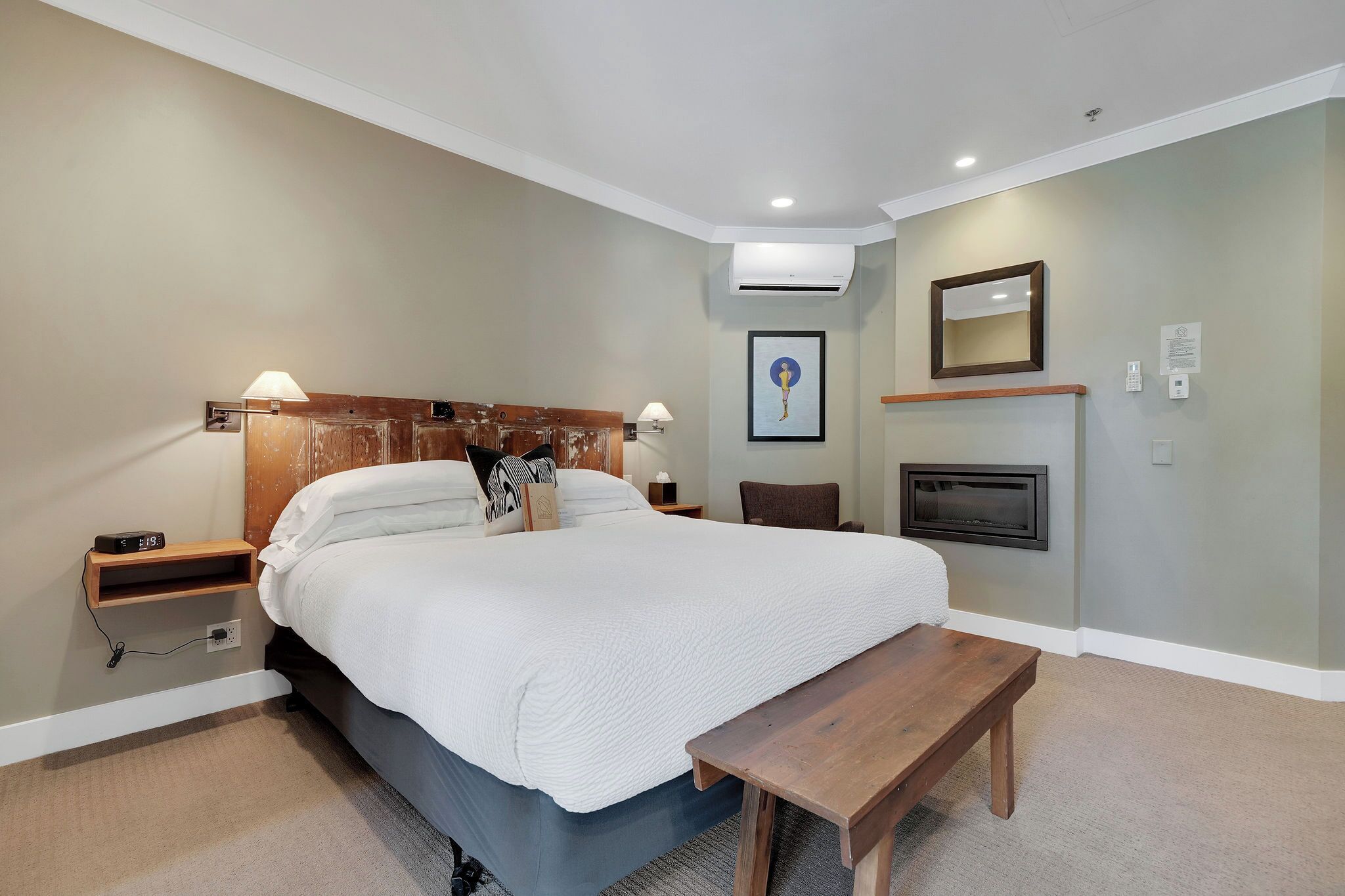 A bedroom with a bed, bedside tables, a bench, an air conditioning unit, a fireplace, and a framed picture on the wall.