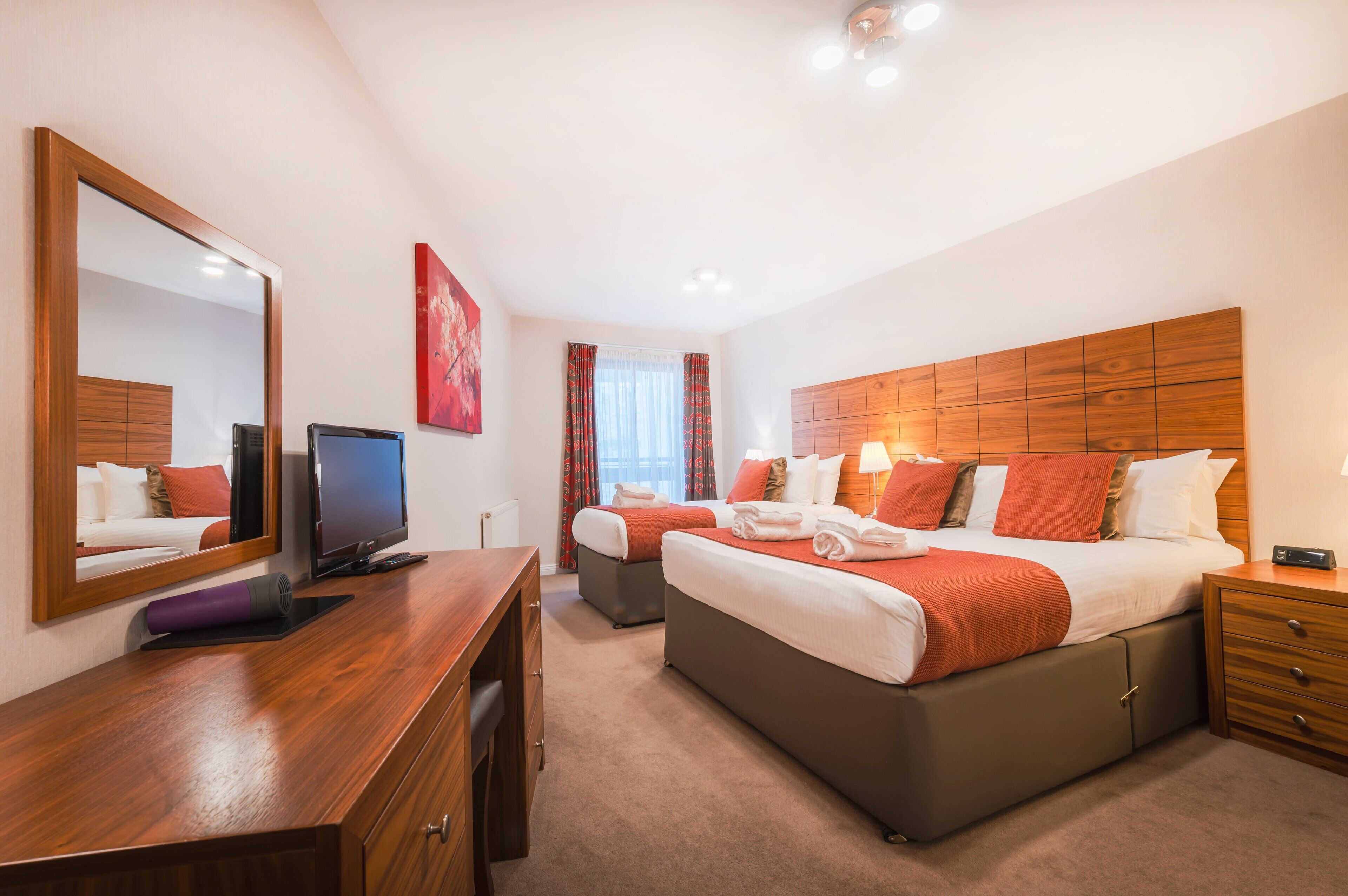 A hotel room with a large bed, a smaller bed, a desk, a TV, and a mirror.