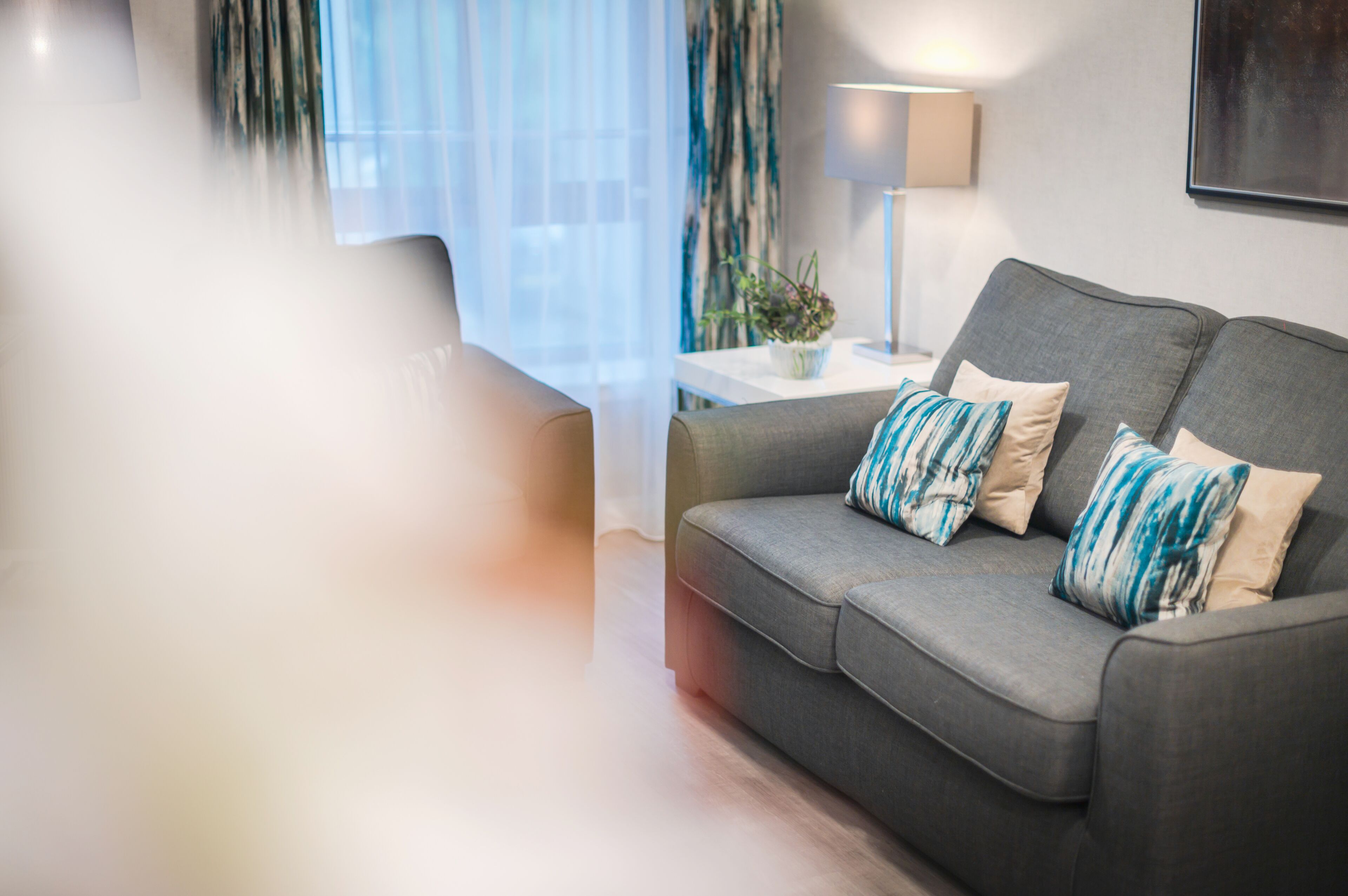 A modern living room with a grey sofa, two patterned cushions, a side table with a lamp, and a window with sheer curtains.