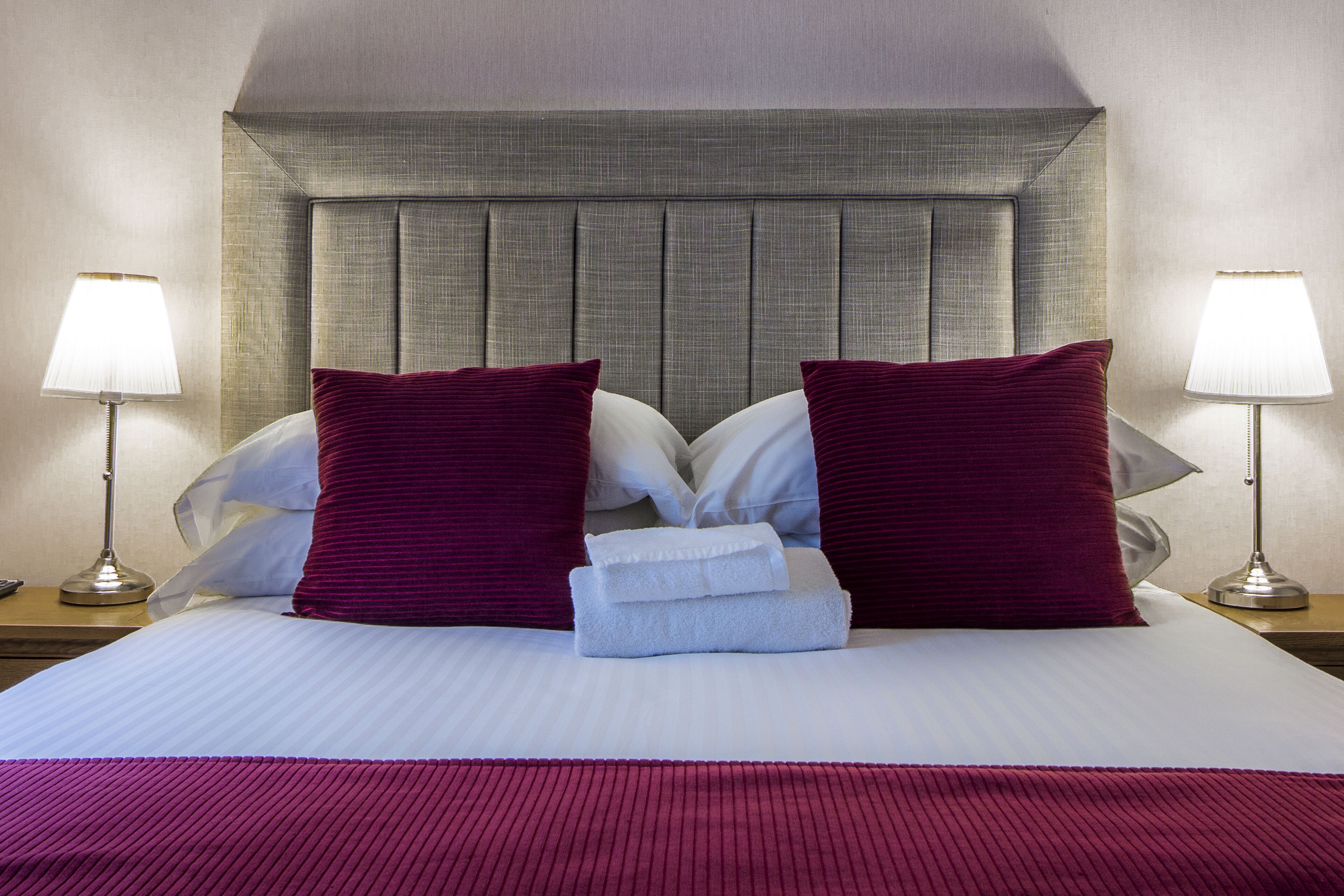 Una cama bien hecha con cabecera, dos almohadas y una toalla doblada. Hay dos lámparas de noche a cada lado de la cama.