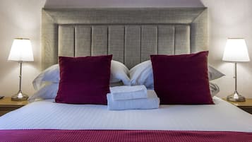 Una cama bien hecha con cabecera, dos almohadas y una toalla doblada. Hay dos lámparas de noche a cada lado de la cama.