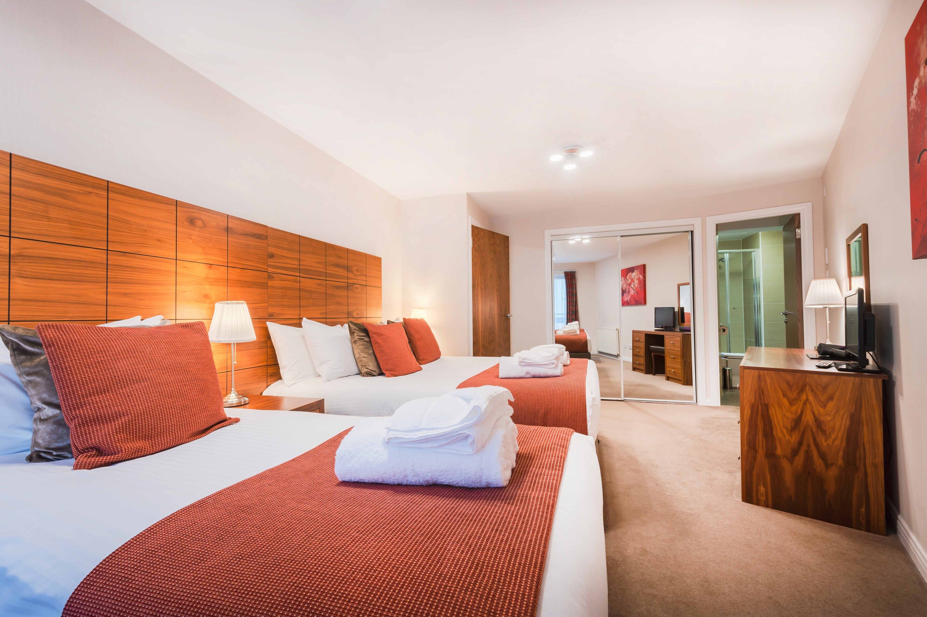 A hotel room with two beds, a wooden headboard, a desk with a television, and a bathroom visible through an open door.