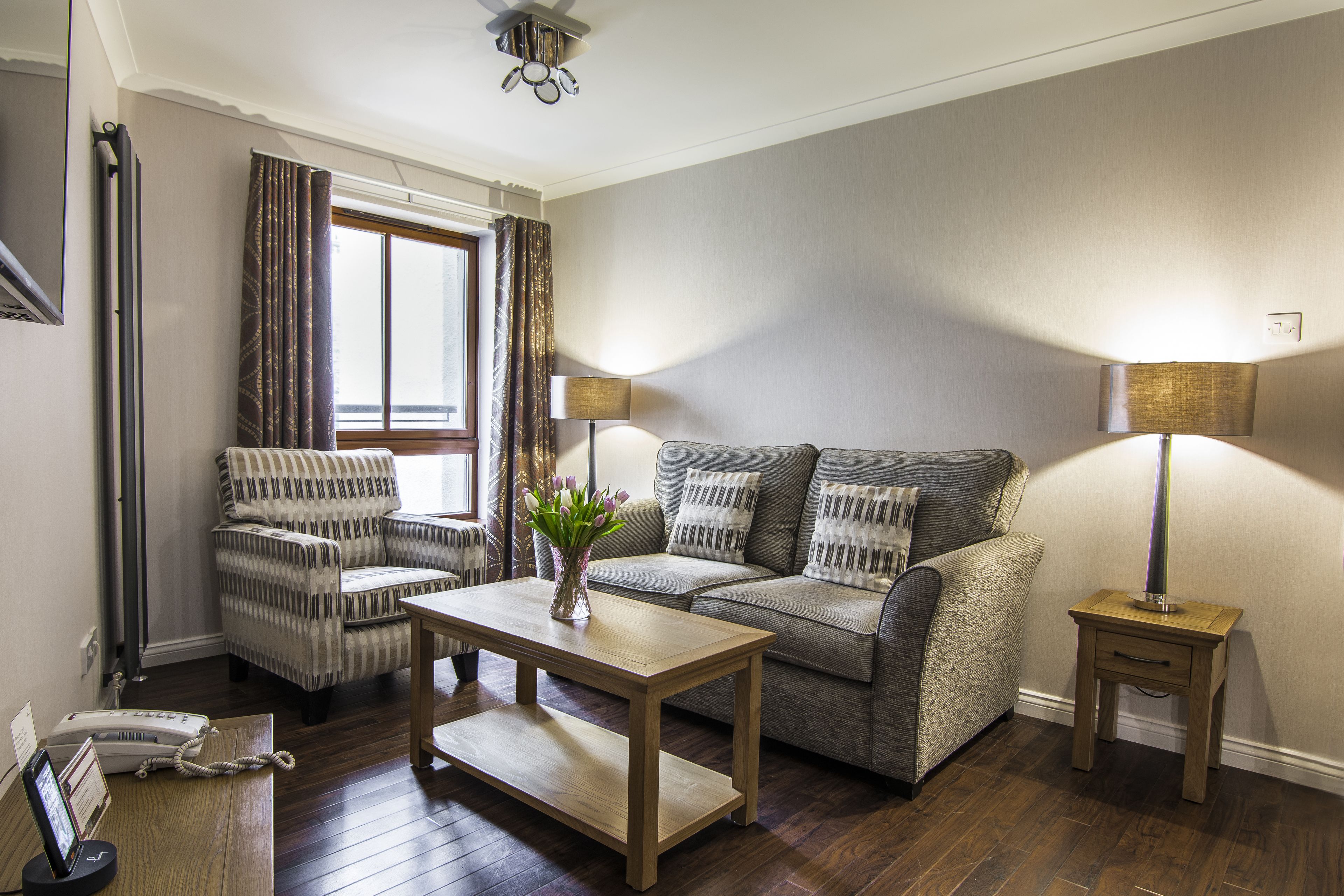 A living room with a sofa, armchair, and a coffee table. There is a lamp on a side table and a window with curtains.