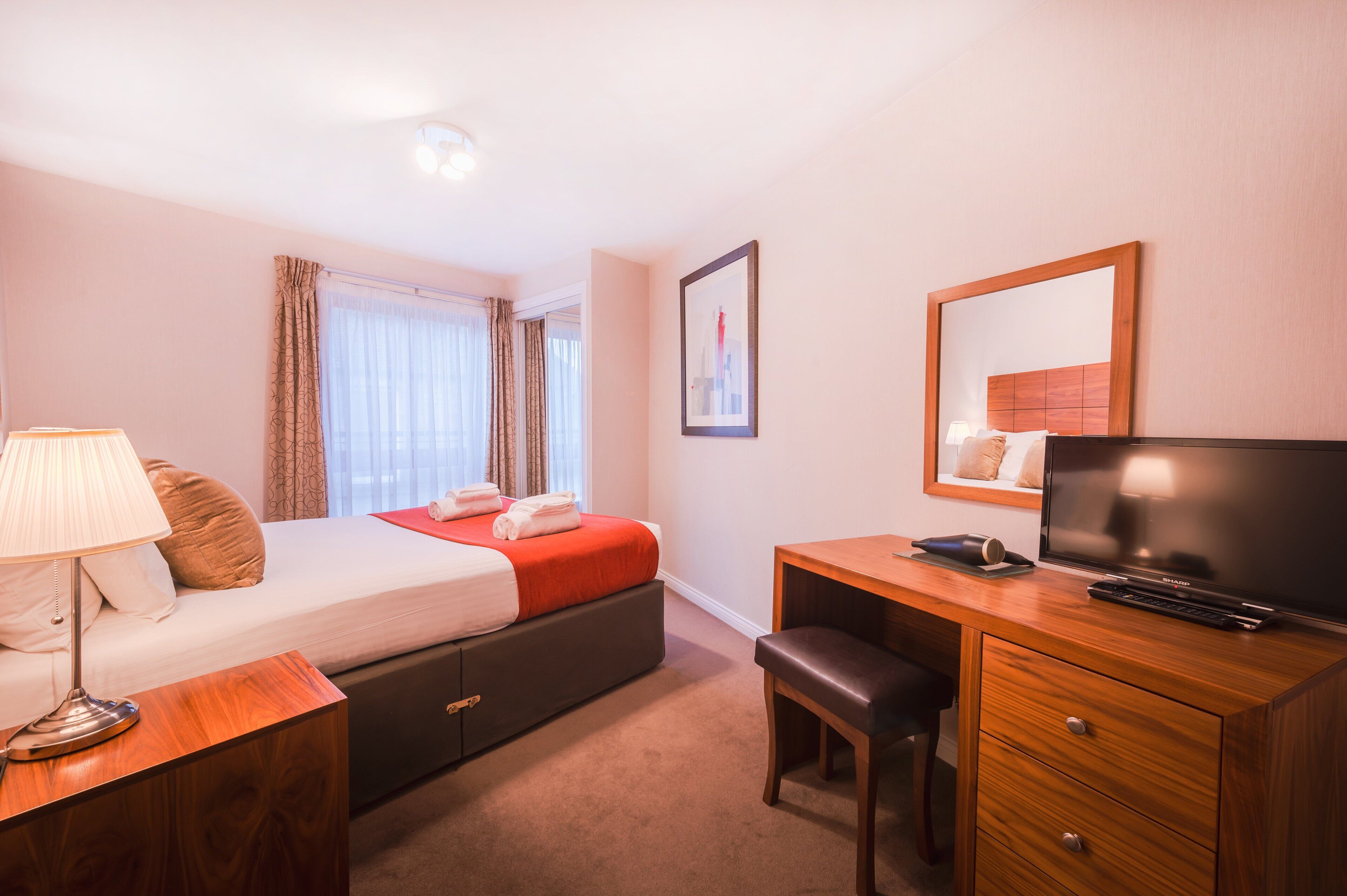 Een hotelkamer met een bed, nachtkastje, lamp, televisie, bureau, stoel en een grote spiegel.