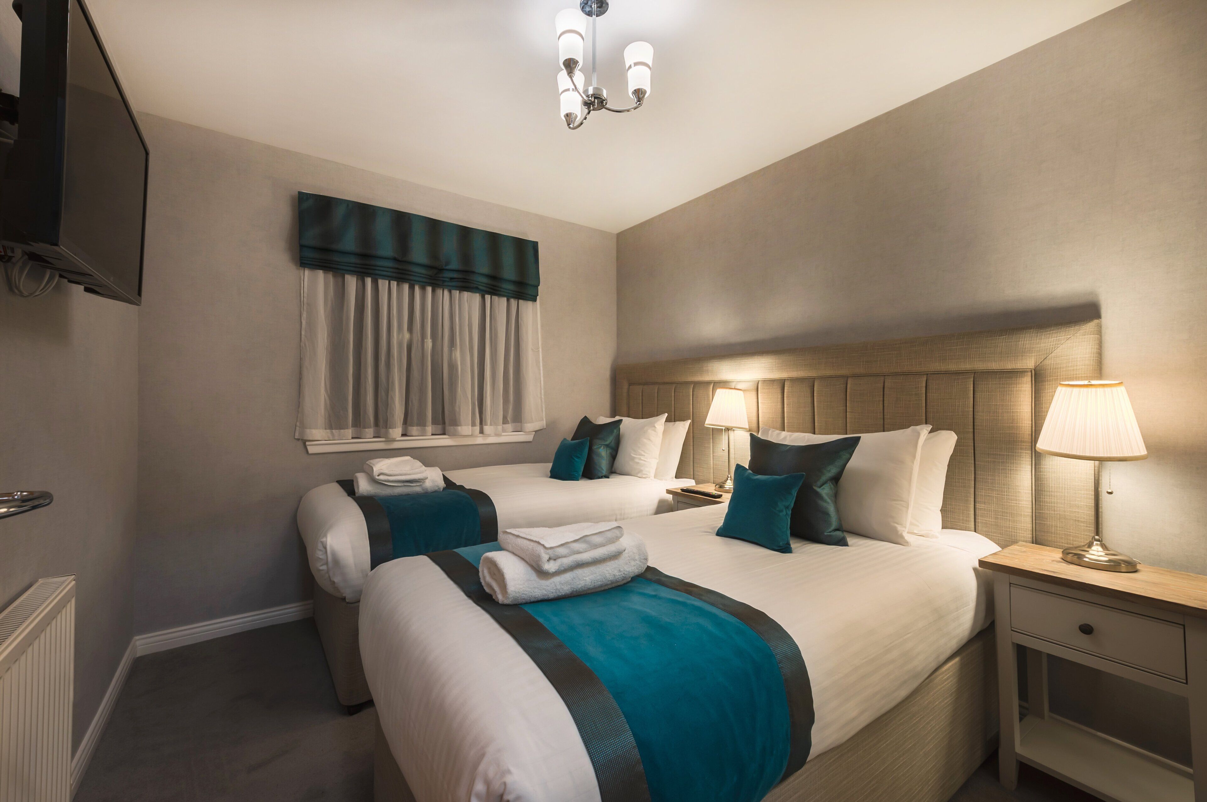 A hotel room with two beds, a TV, a chandelier, and a window with curtains.